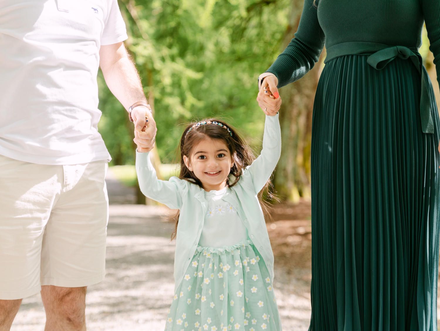 A joyful little girl in a mint dress holds hands with her parents during an outdoor family photo session.