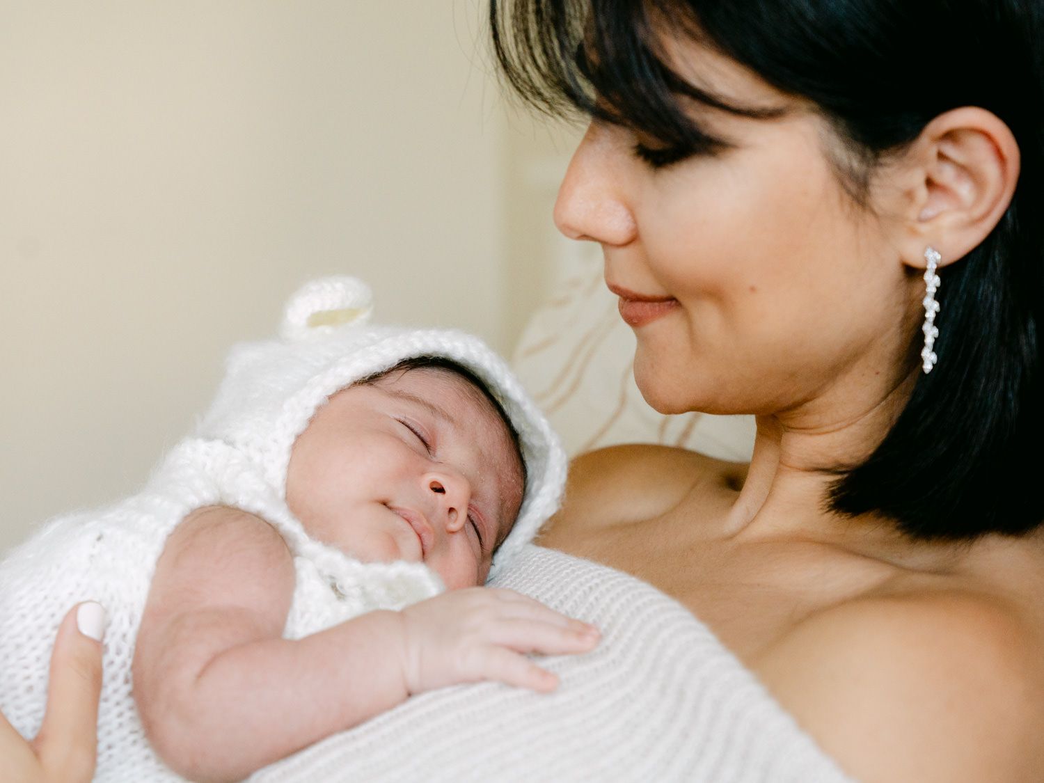 Mother with drop earrings tenderly gazes at sleeping newborn in white bear bonnet outfit.