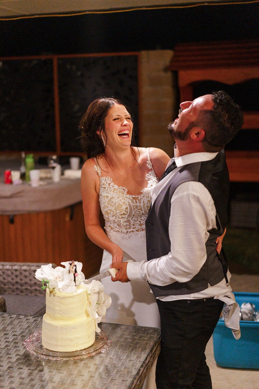 Bride and groom laughing joyfully while cutting their white wedding cake together at an outdoor evening reception.