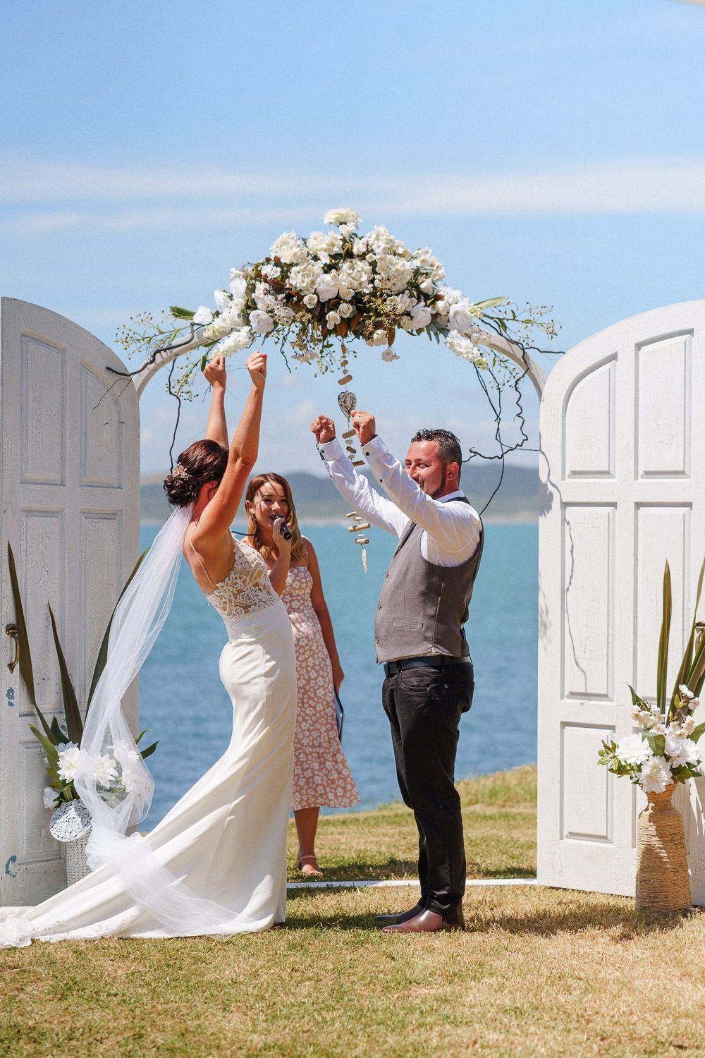 Bride and groom celebrating under floral arch by the sea.