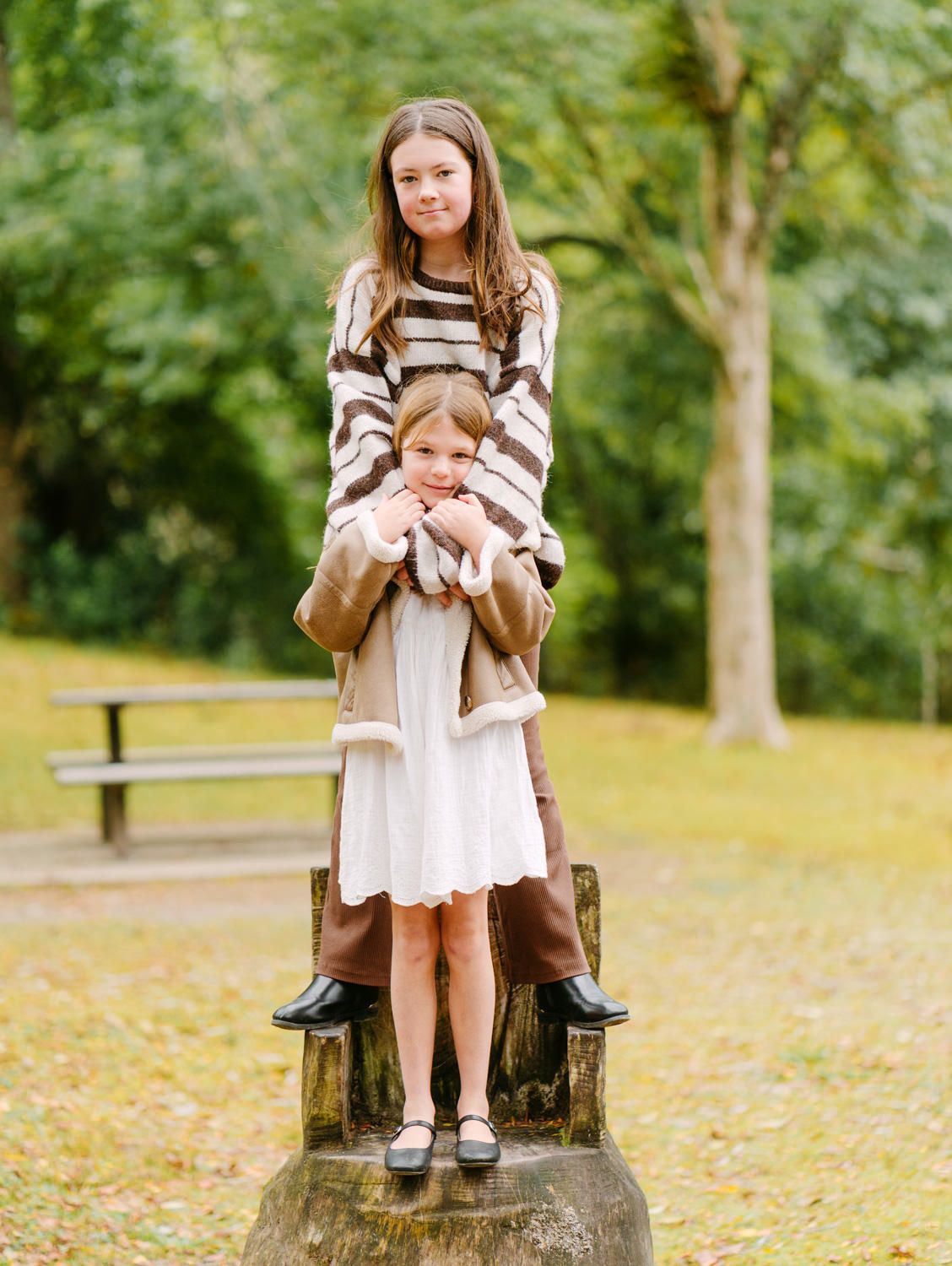Two girls pose on a tree stump in a park, wearing autumn outfits, surrounded by green and yellow foliage.