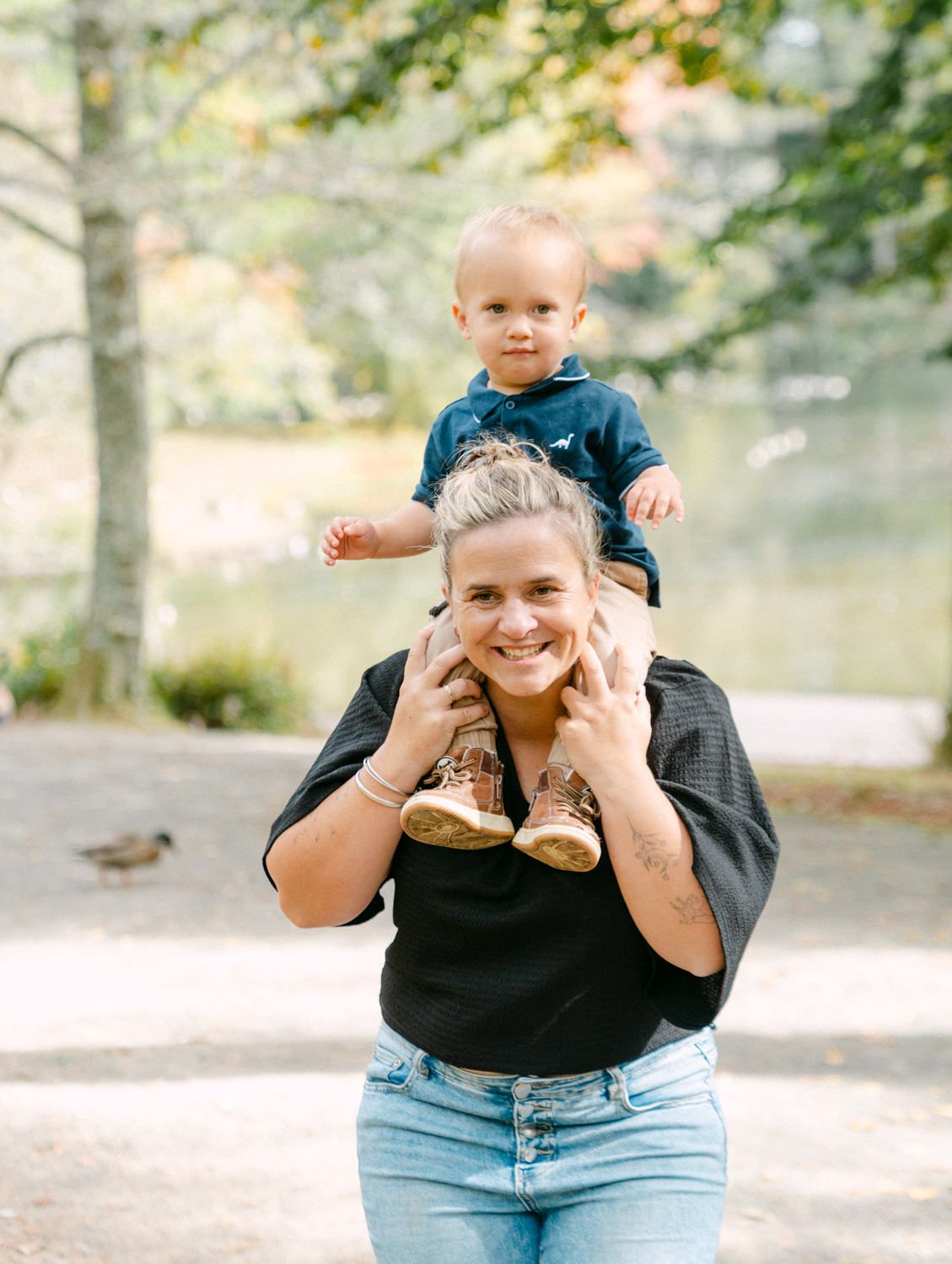 A smiling woman carries a happy toddler on her shoulders in a sunny park setting.