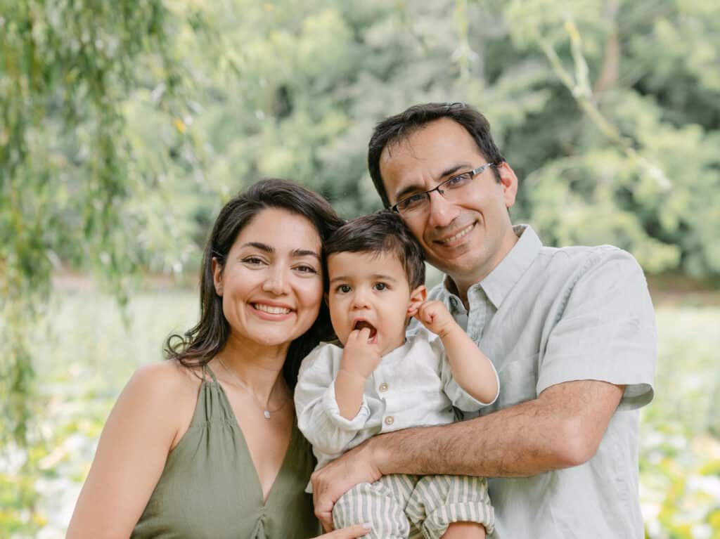 Happy family portrait of smiling parents holding their baby outdoors with lush green trees in background.