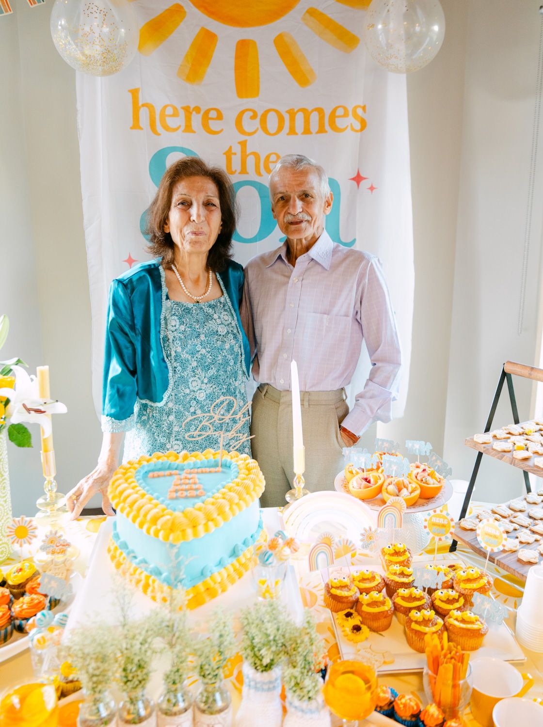Couple posing behind a dessert table with heart cake and cupcakes at a sunshine-themed celebration party.