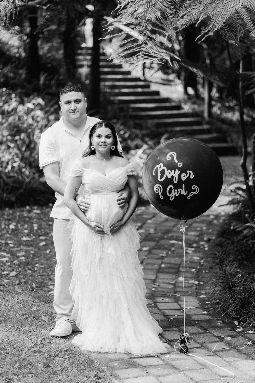 Pregnant couple poses with boy or girl gender reveal balloon in black and white outdoor maternity photo.