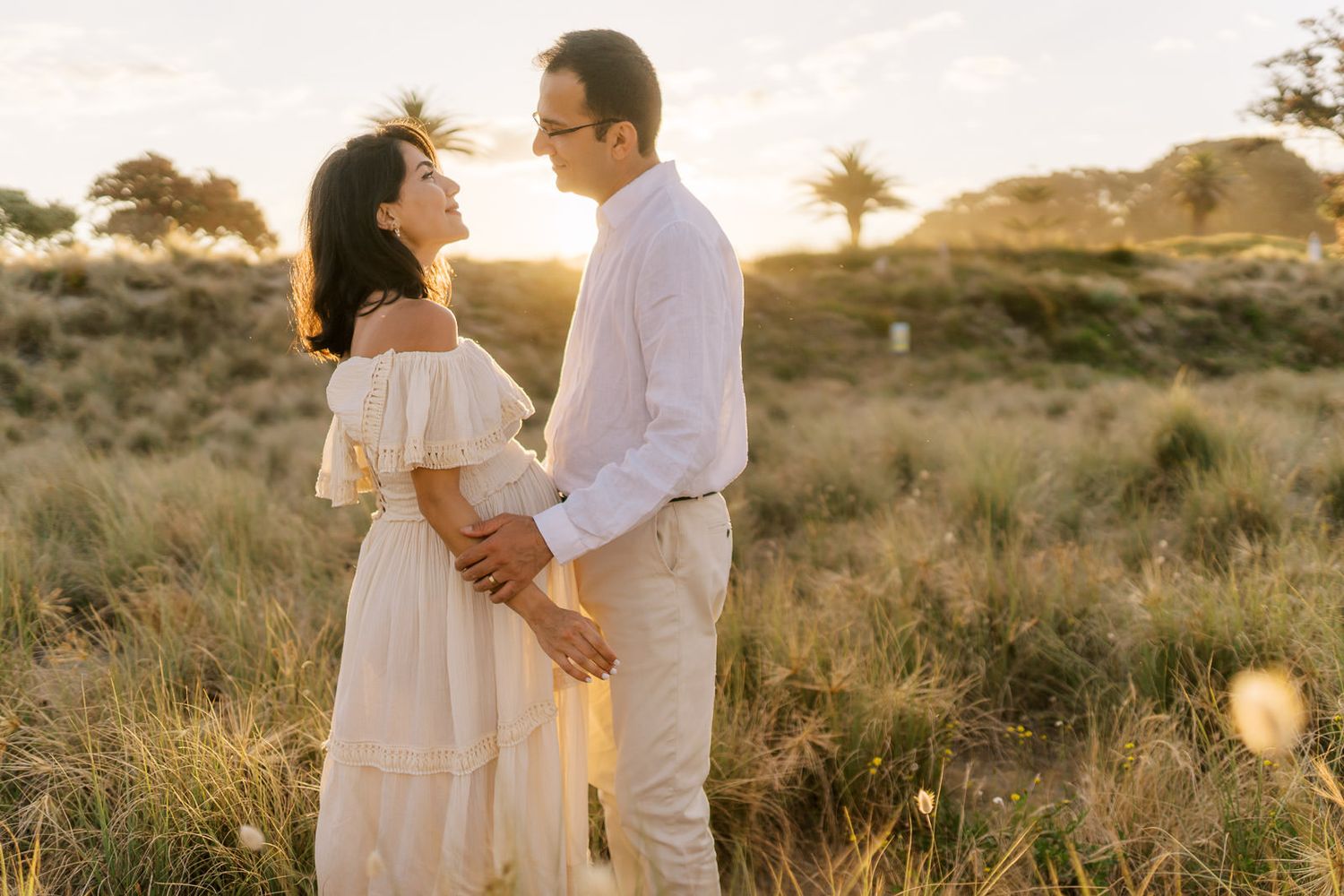 Couple in white outfits embracing in golden sunset field, romantic outdoor maternity photo session.