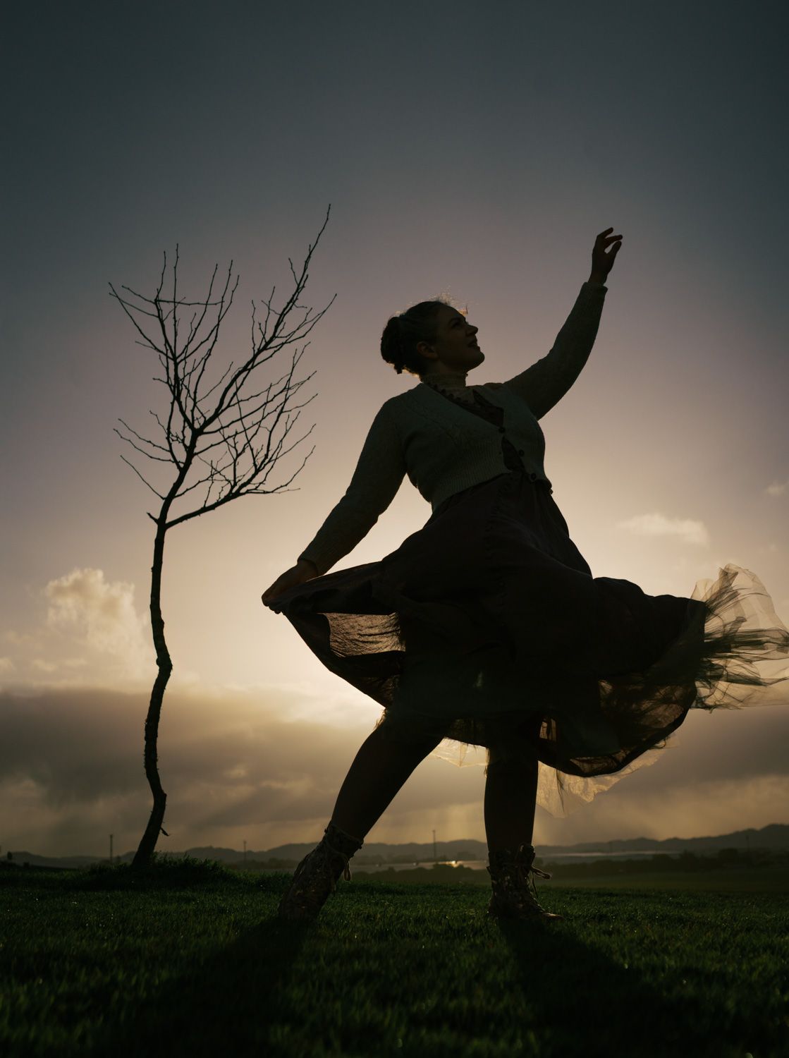 Silhouette of a woman dancing in a flowing skirt at sunset near a bare tree in an open field.