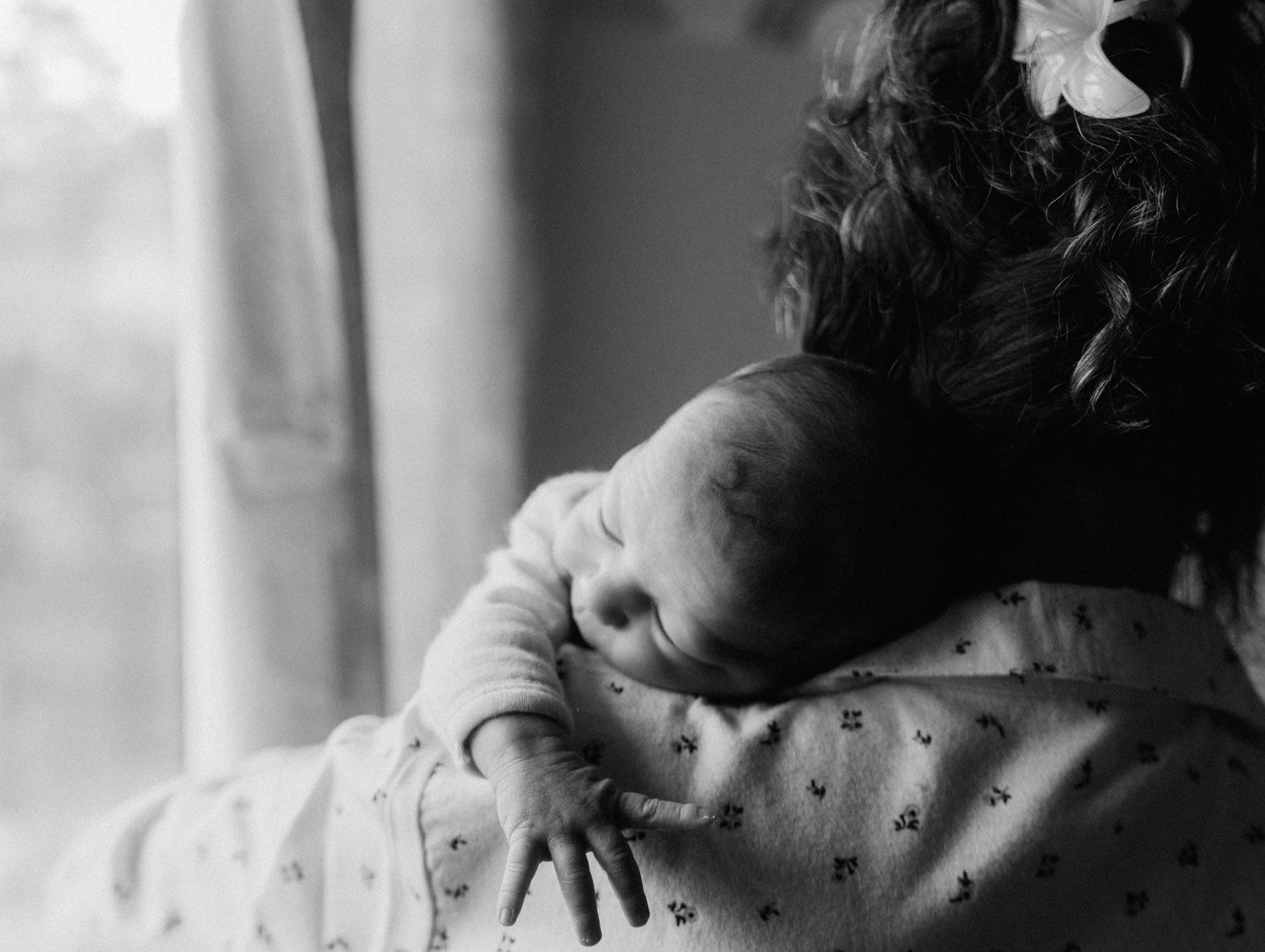 Black and white photo of a mother cradling a newborn baby on her shoulder near a window.
