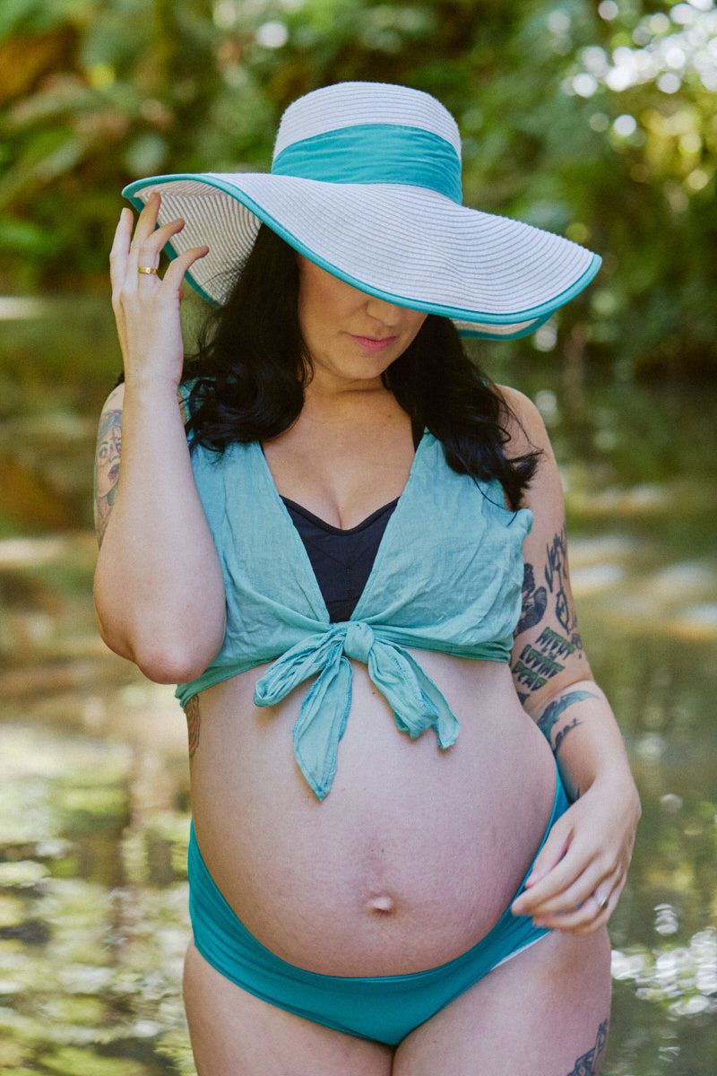 Pregnant woman in teal bikini and wide-brim sun hat standing in water surrounded by lush tropical greenery.