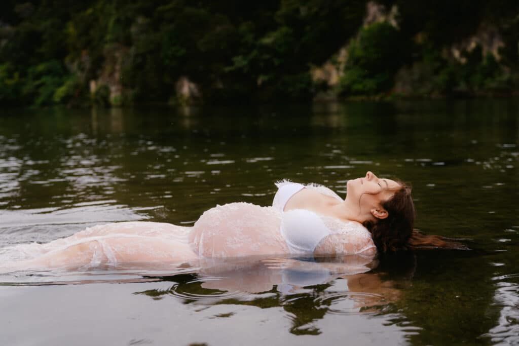 Woman in white dress floating peacefully in a calm lake surrounded by lush green trees.