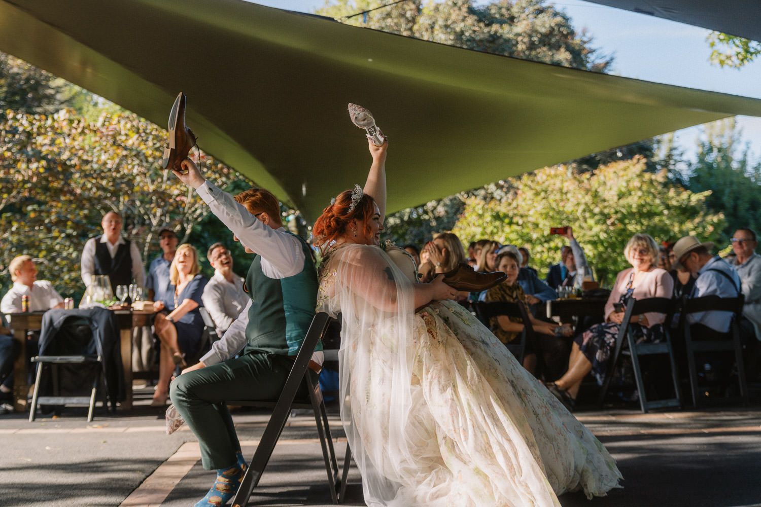 Bride and groom dancing joyfully outdoors at wedding reception under green canopy with guests seated in background.