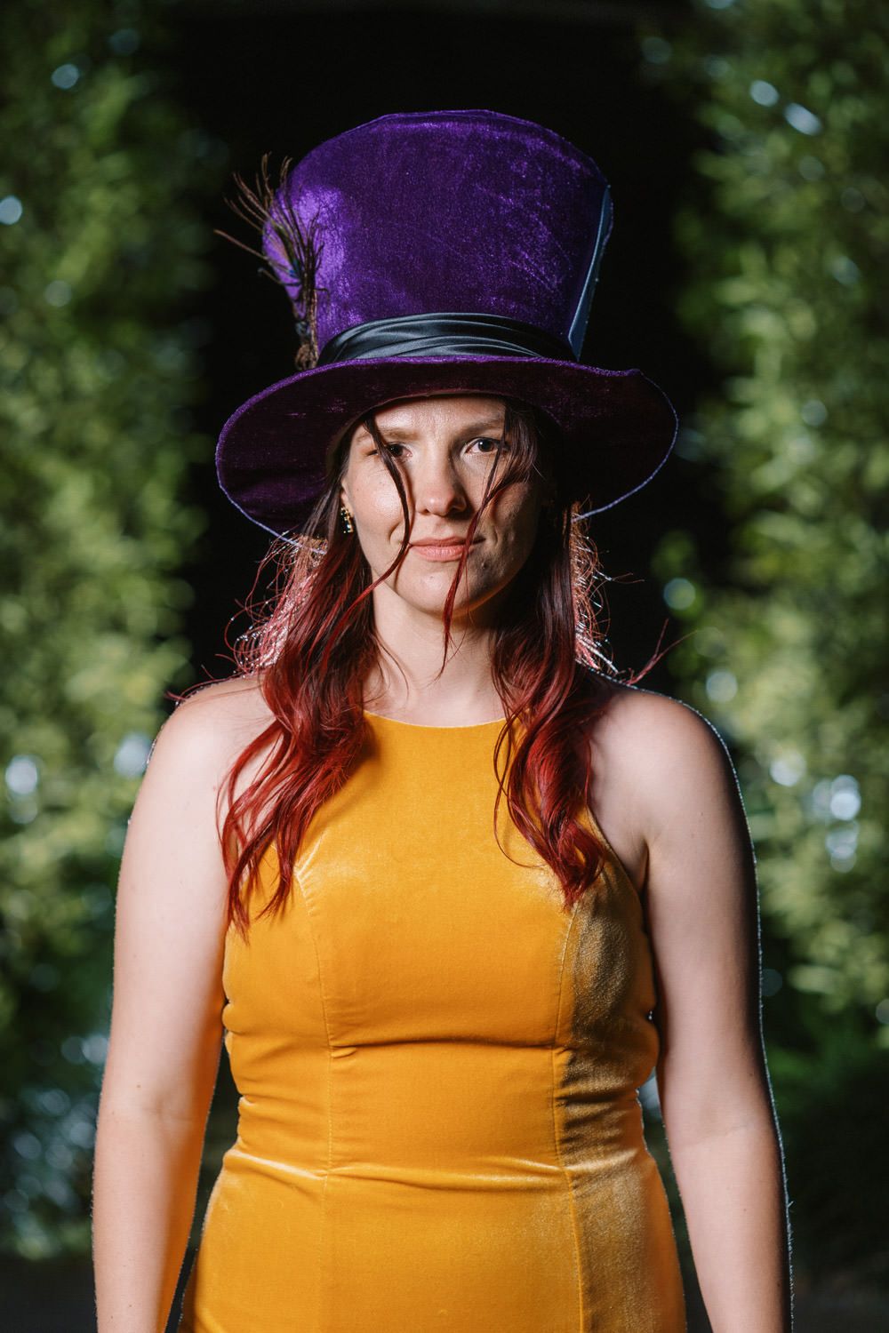 Woman with red hair wearing a purple velvet top hat and yellow dress poses outdoors with green foliage background.