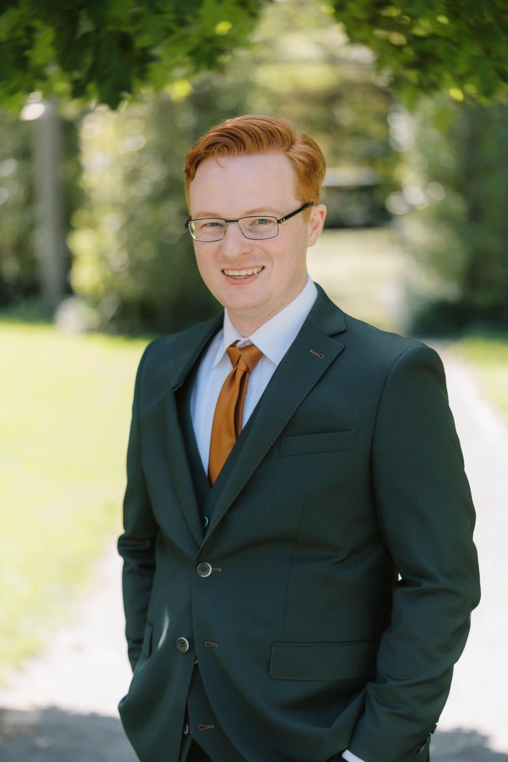 Young redhead man in green suit, orange tie, and glasses smiling outdoors in a sunny park setting.
