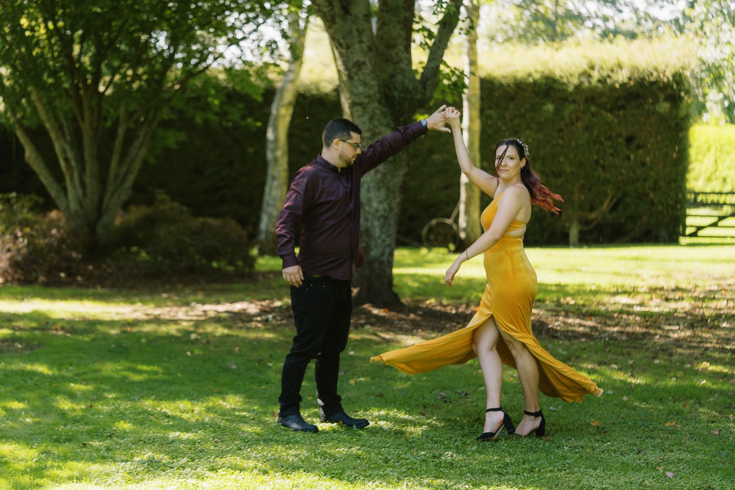 Couple dancing outdoors in a lush green garden, woman in flowing yellow dress, man in dark maroon shirt.