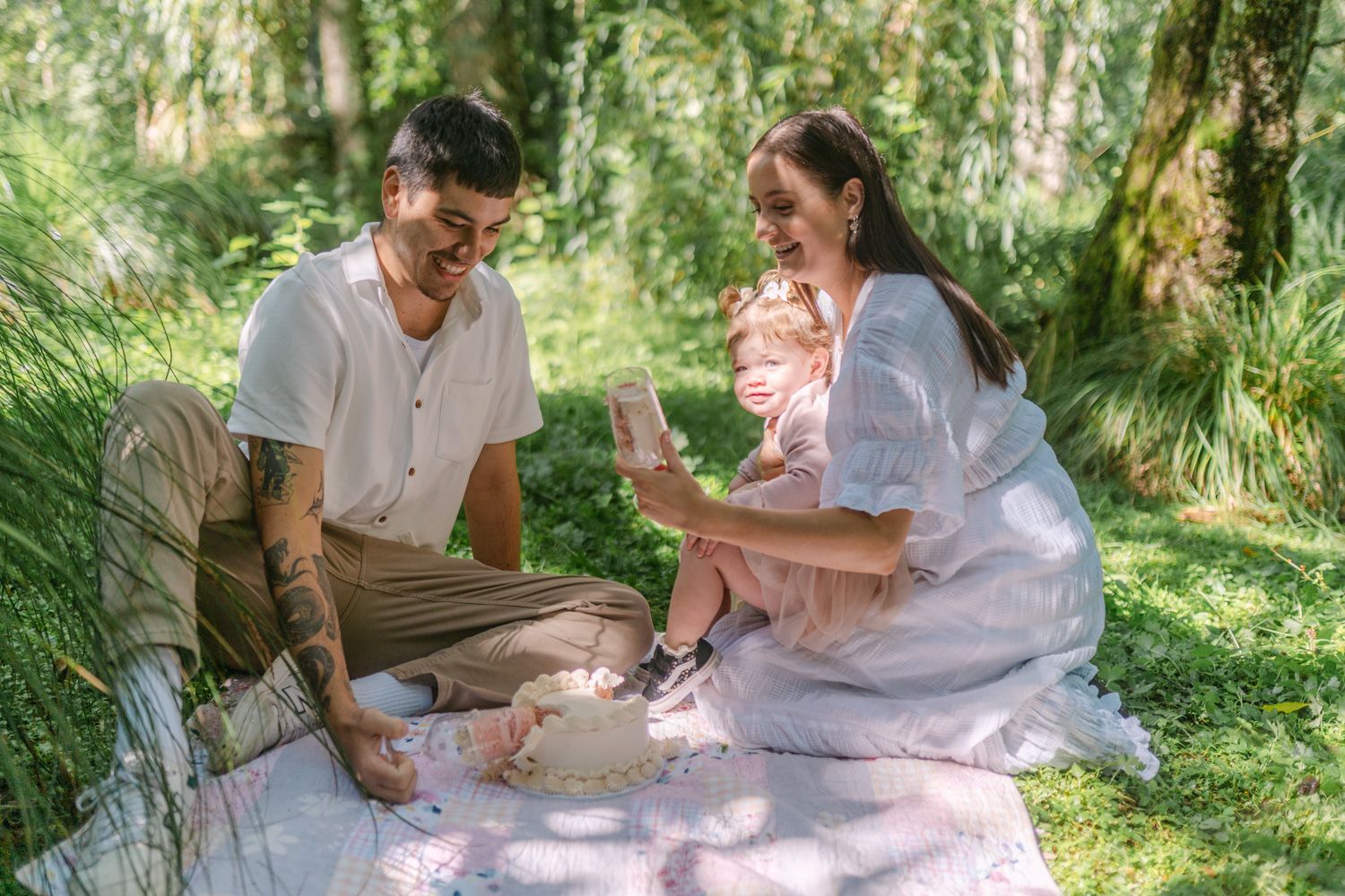 A young family enjoys a sunny outdoor picnic on a blanket surrounded by lush green trees.