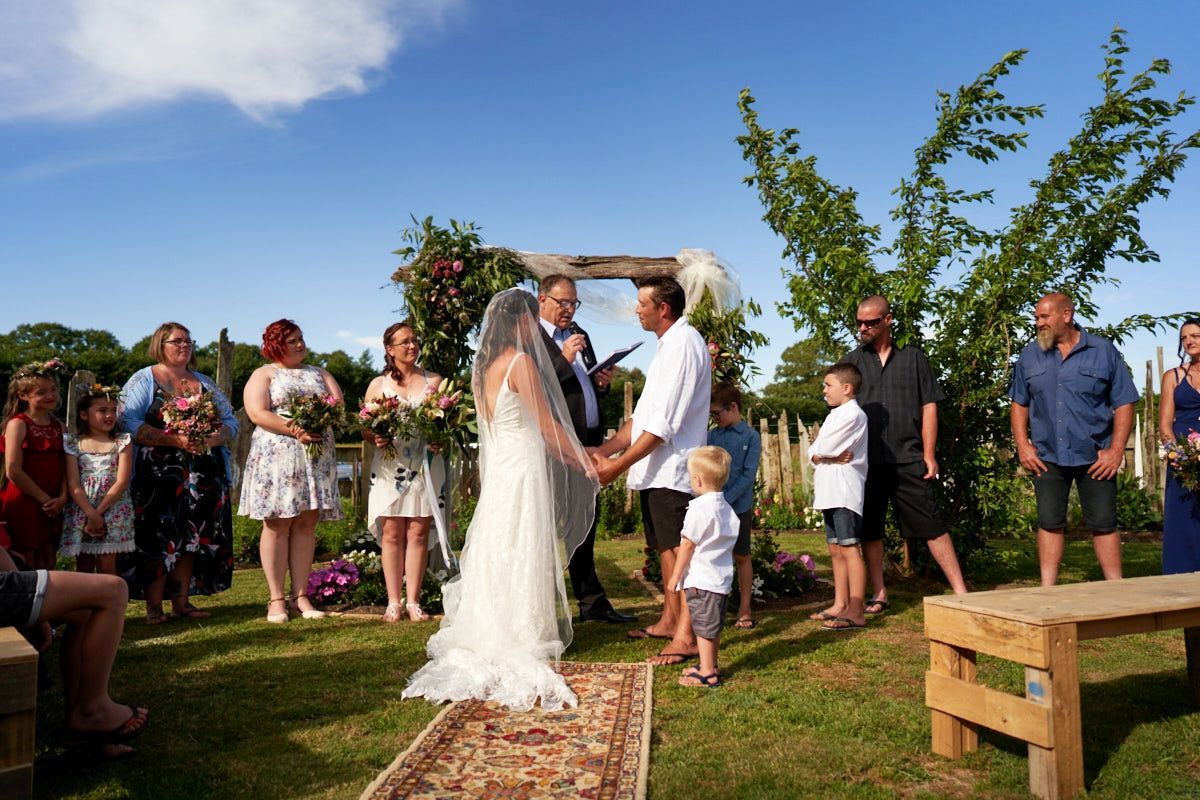 Outdoor wedding ceremony with bride and groom at floral arch, surrounded by guests on a sunny day.