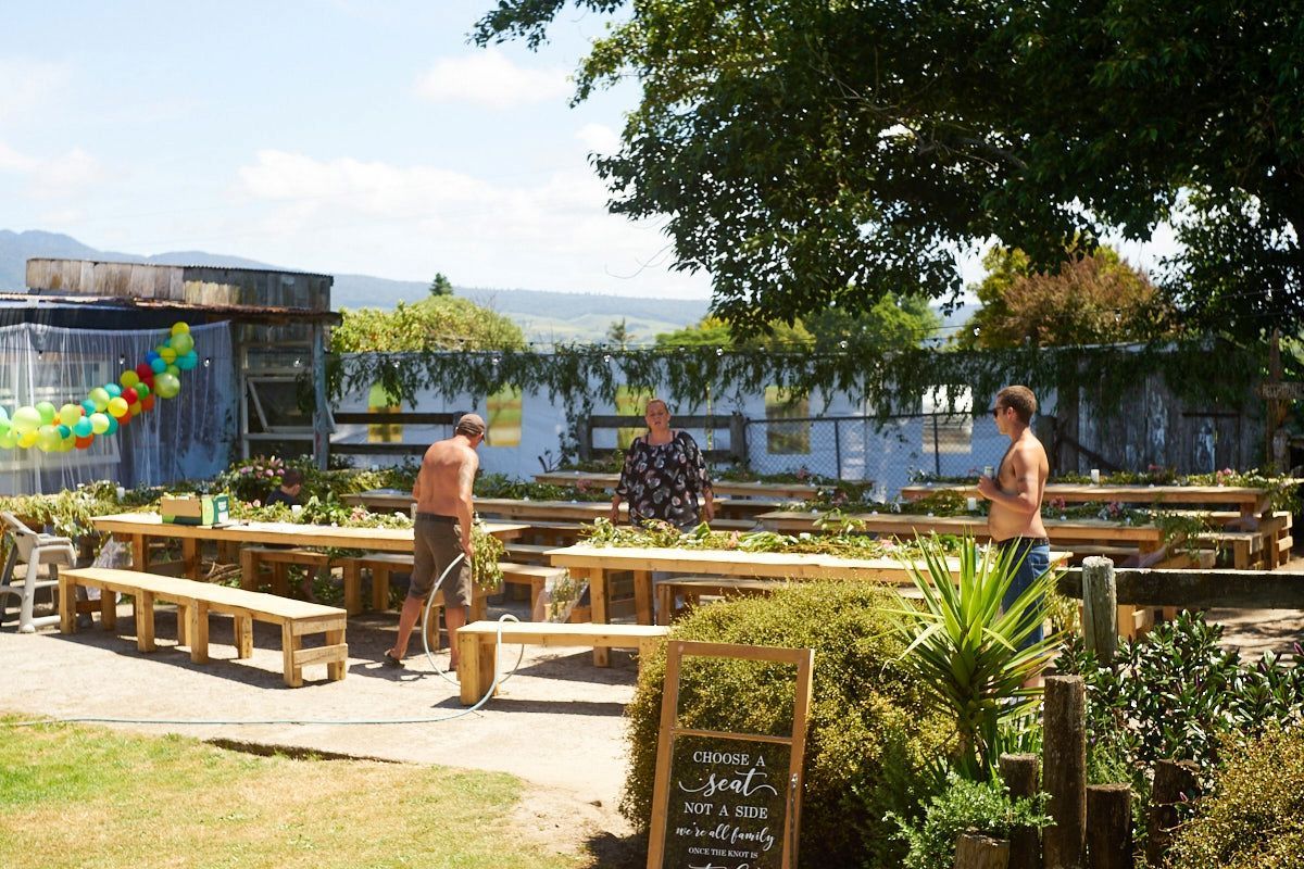 Outdoor garden seating area with wooden benches and tables, people socializing under a sunny sky near lush greenery.