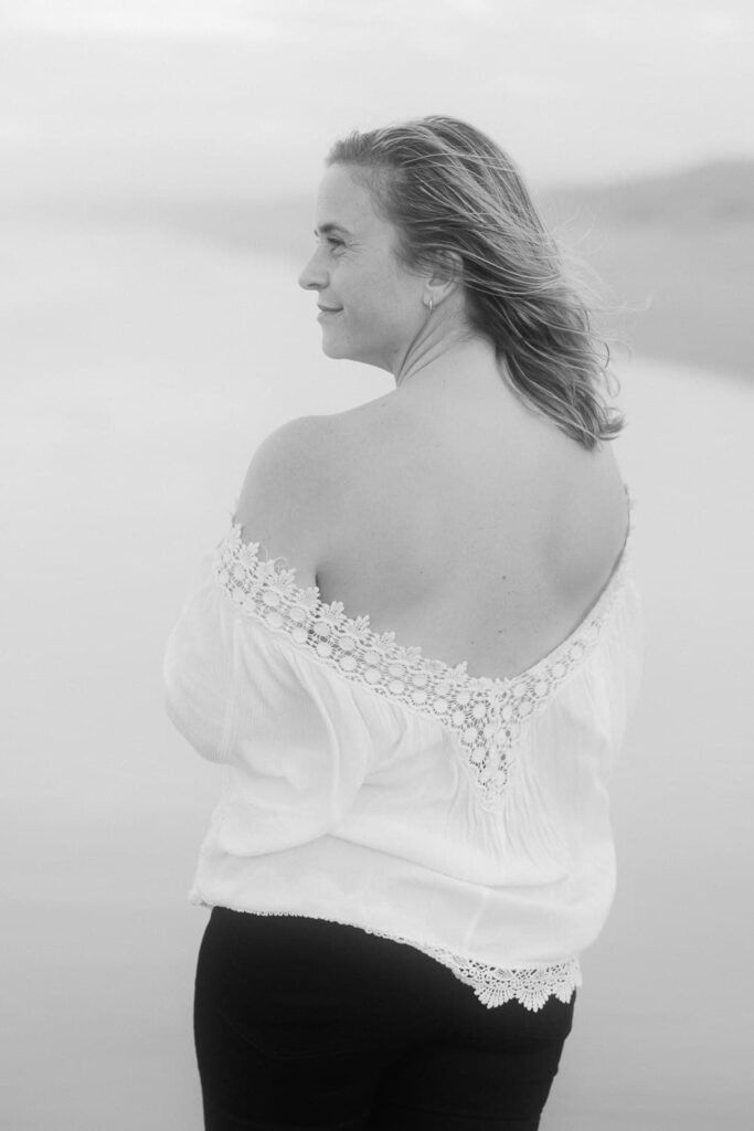 Black and white photo of a woman in an off-shoulder white lace top, looking away at a serene beach backdrop.