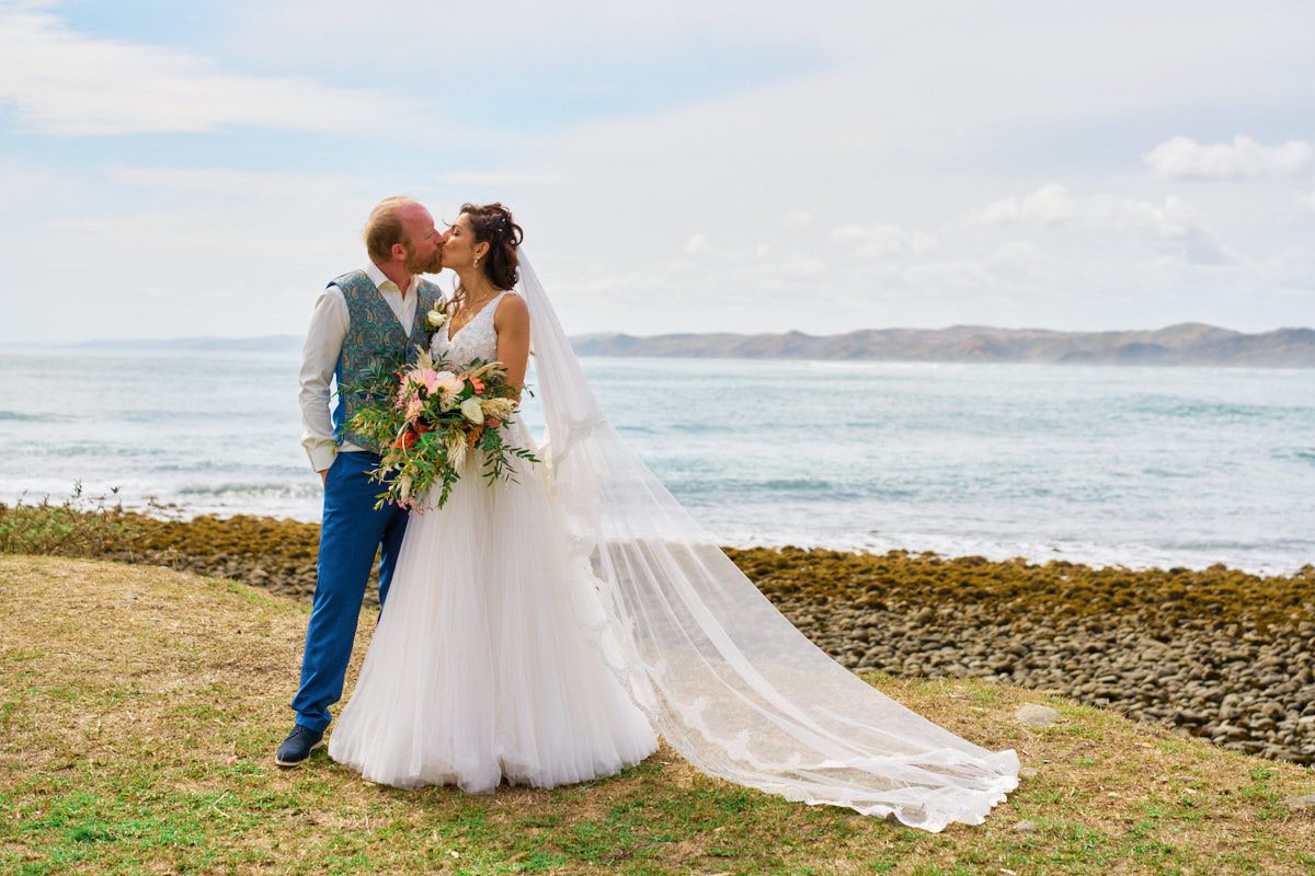 Bride and groom share a kiss by the ocean, with rolling waves and green hills in the background on their wedding day.