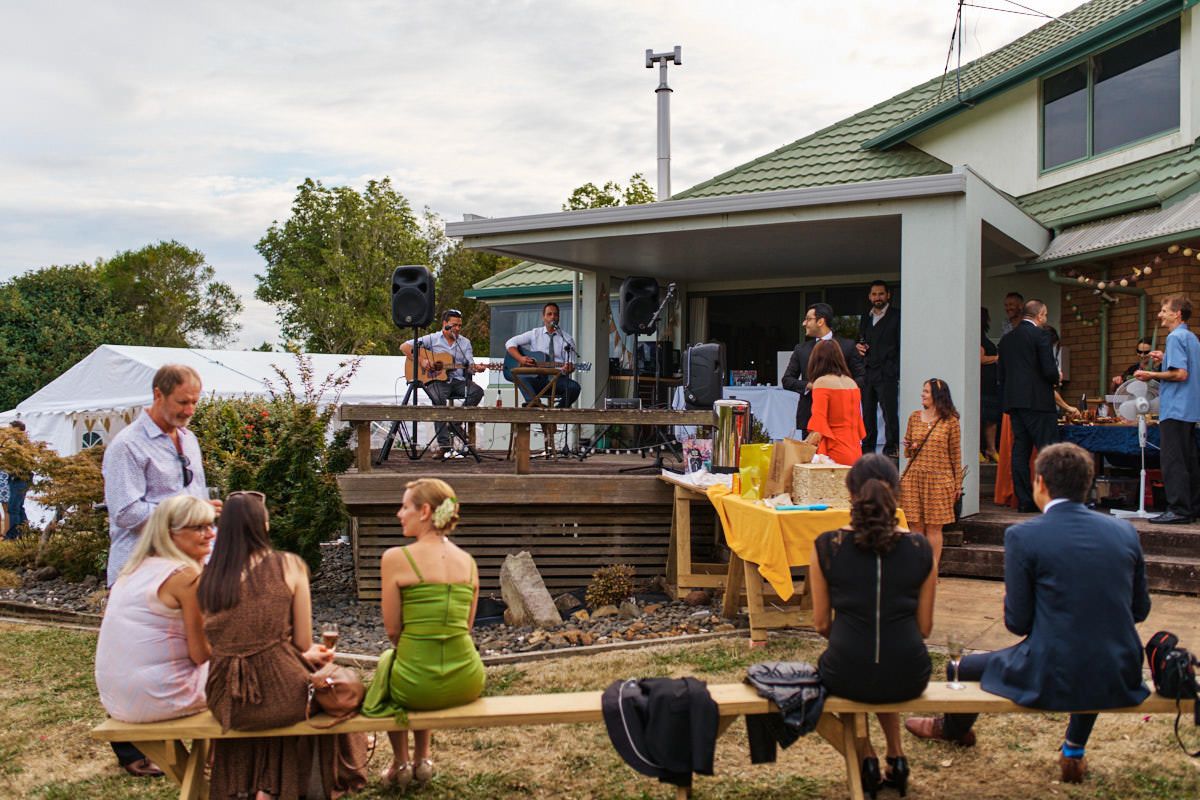 Outdoor backyard party with live band performing on deck while guests socialize on wooden benches.