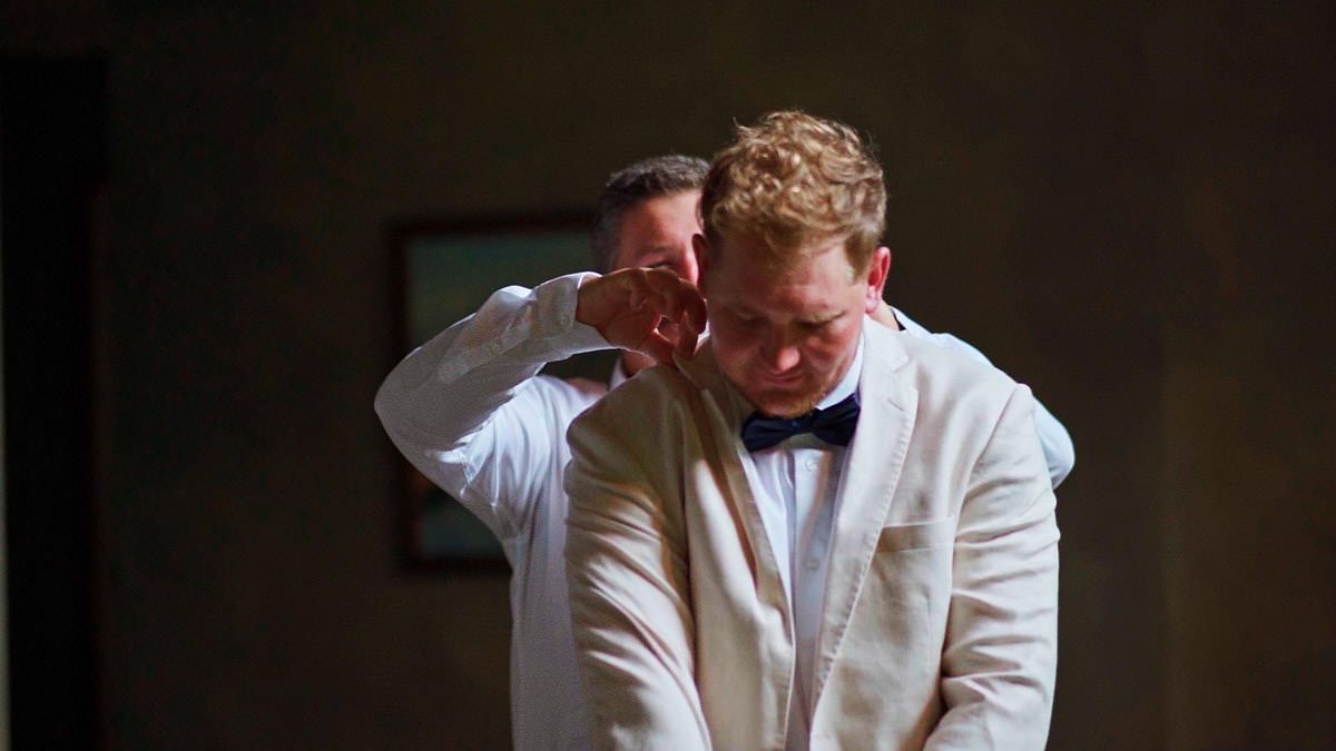 Two men in white suits, one adjusting the other's bow tie in an elegant indoor setting.