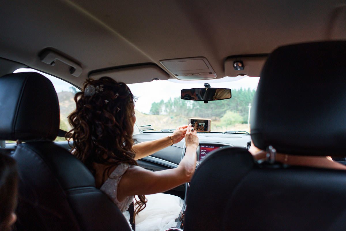 Bride in wedding dress adjusting rearview mirror while sitting in car passenger seat.