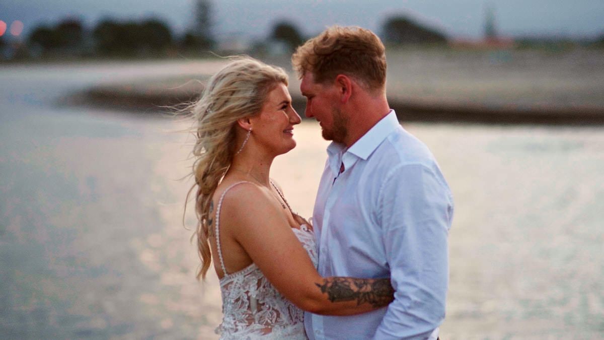 A romantic couple embraces near a shimmering waterfront at sunset, with the woman's blonde hair blowing in the breeze.