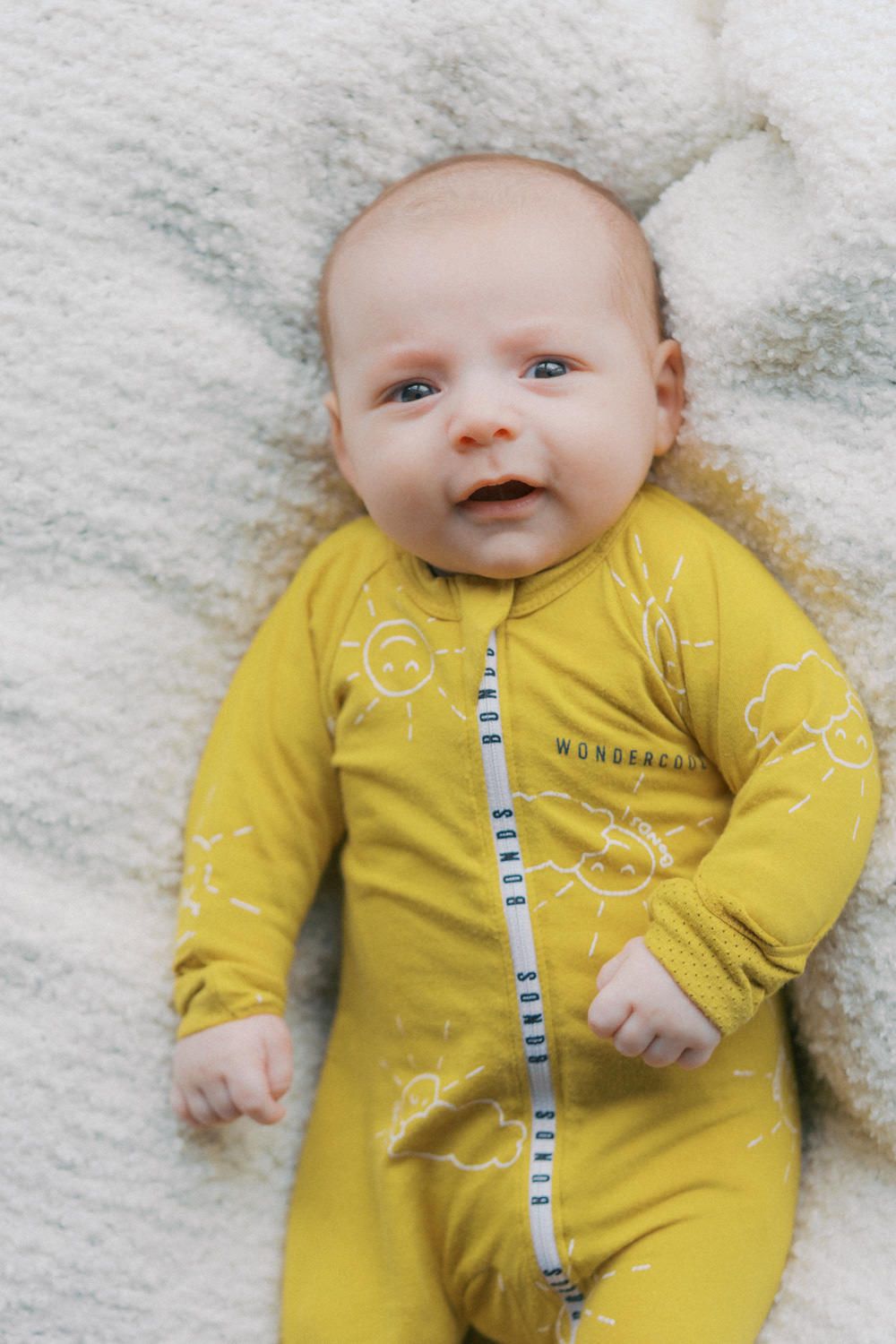 Baby in yellow Bonds Wondercool zipsuit lying on white fluffy blanket, looking up with bright blue eyes.
