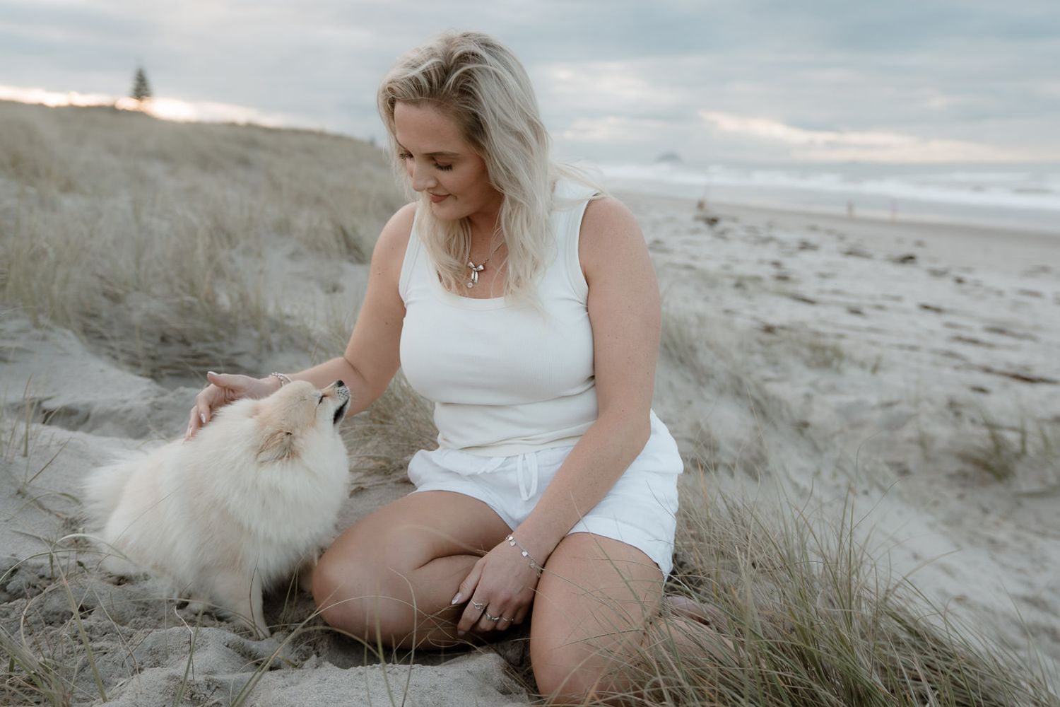 Woman in white outfit sits on sandy beach dunes petting her fluffy white Pomeranian dog near the ocean.