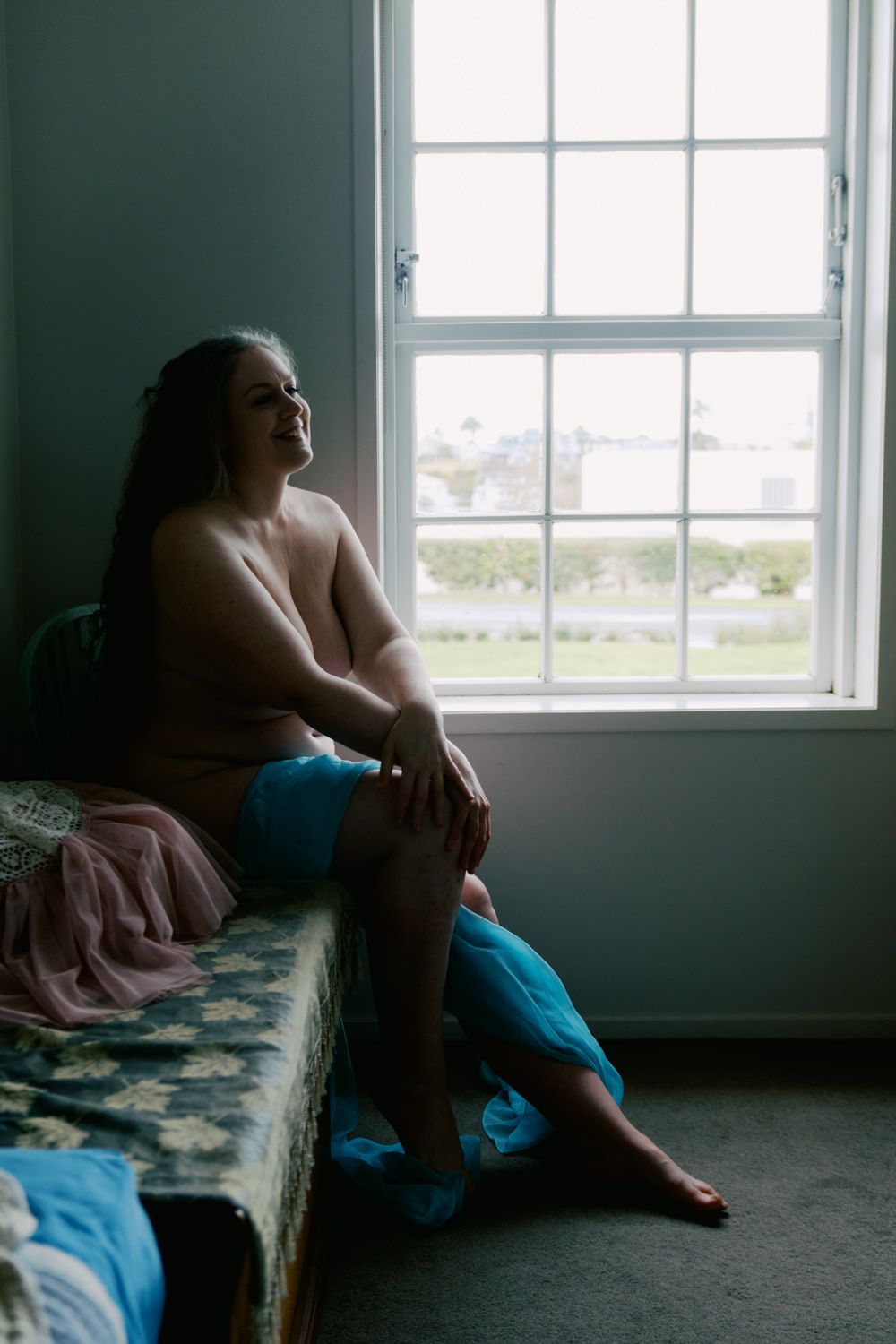 Person sitting on a bed by a bright window, gazing upward in a contemplative mood, wearing blue fabric.
