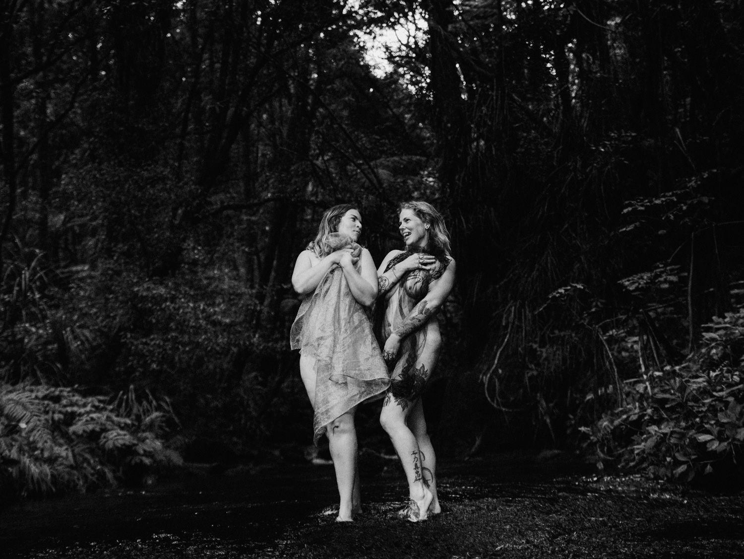 Two women in flowing garments stand together in a black and white forest scene near a stream.