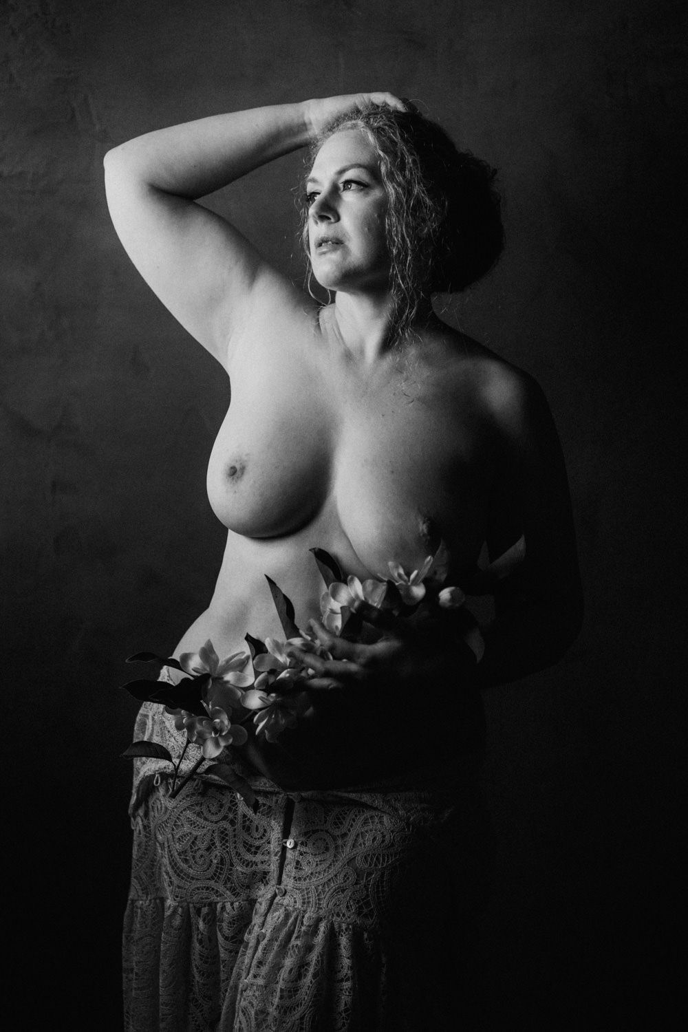 Black and white artistic nude portrait of a woman holding flowers, posed against a dark studio background.