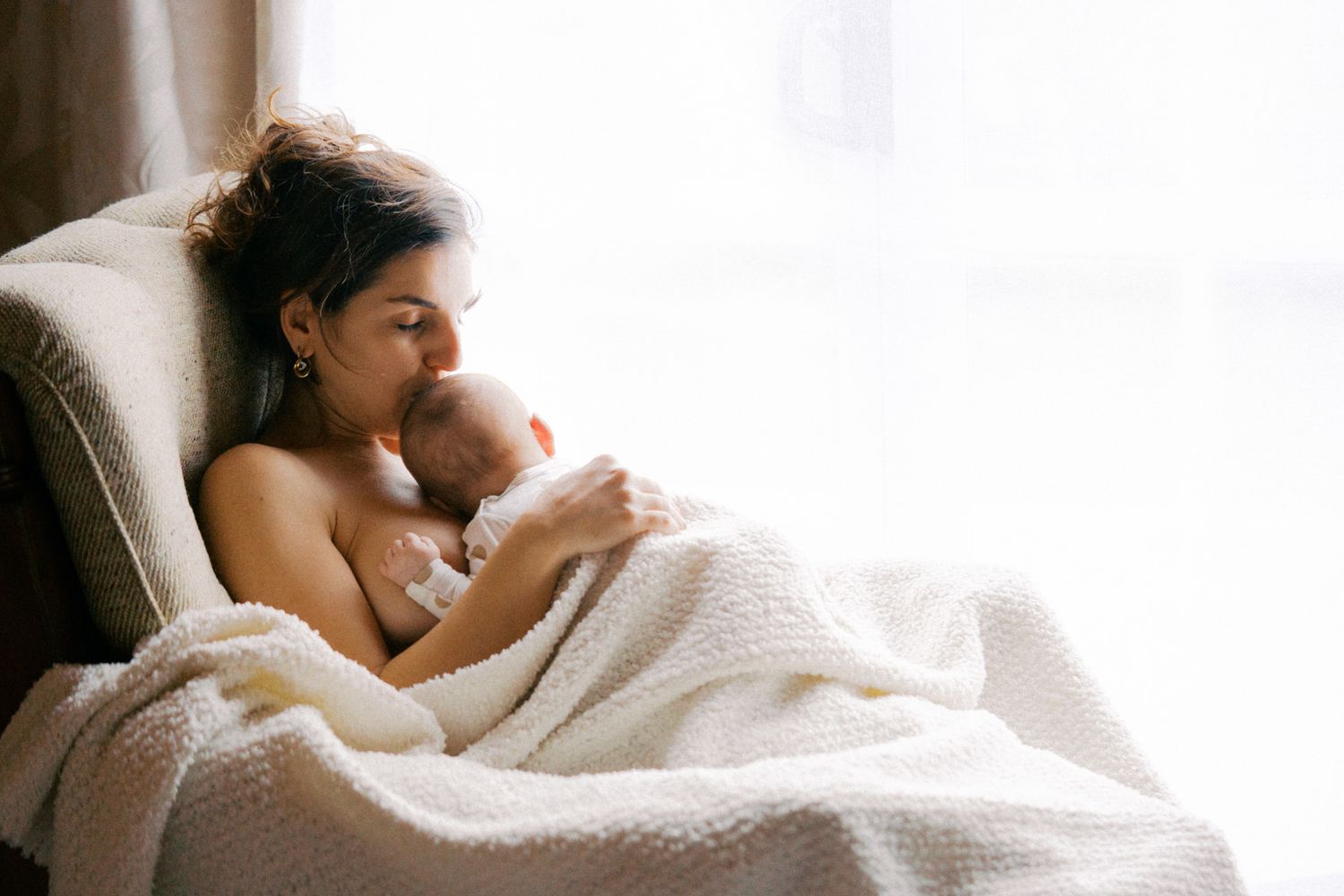 Mother tenderly holding and kissing her newborn baby wrapped in a white blanket while sitting in an armchair.
