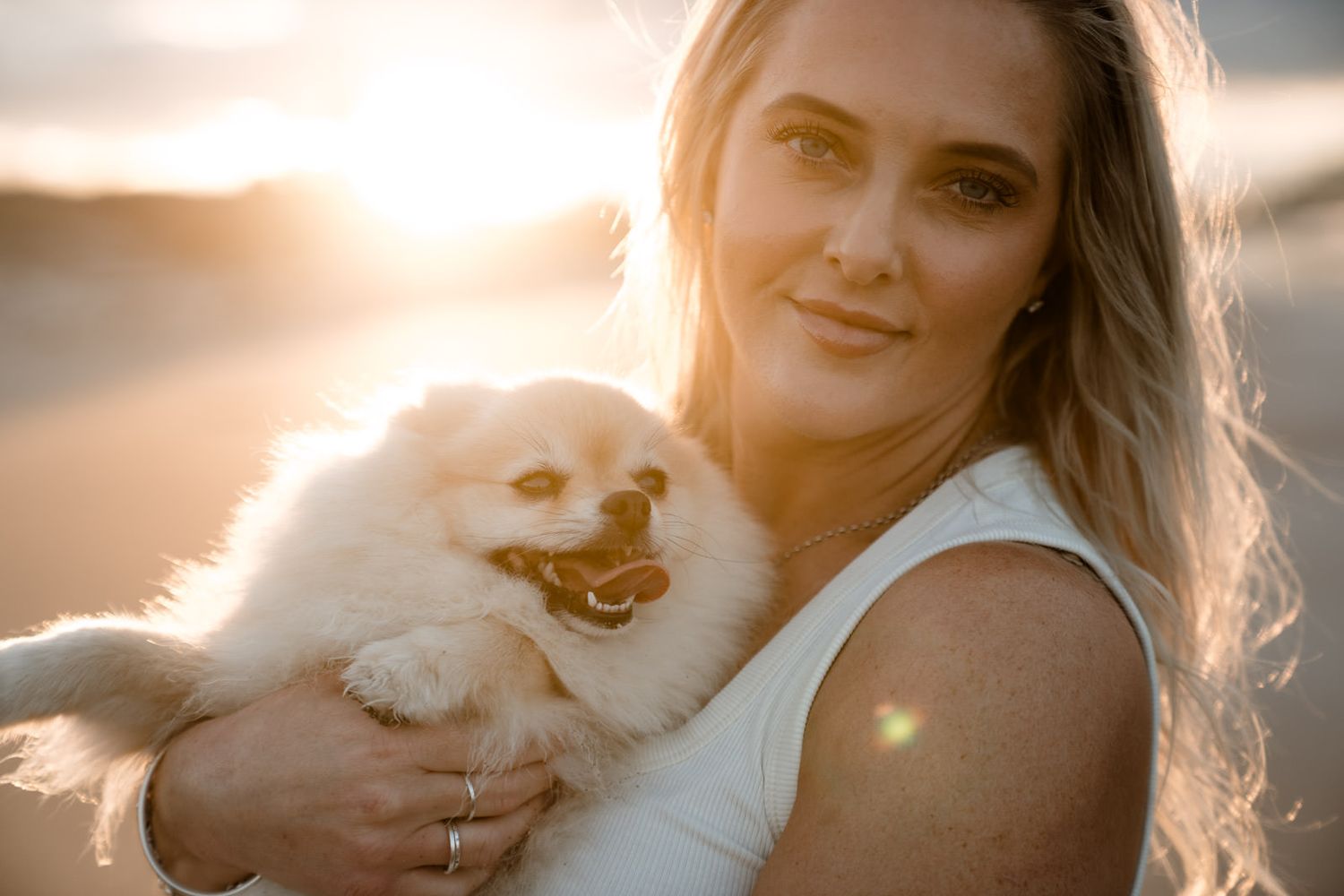 Woman holding fluffy white Pomeranian dog at golden hour beach sunset, warm glowing backlight.
