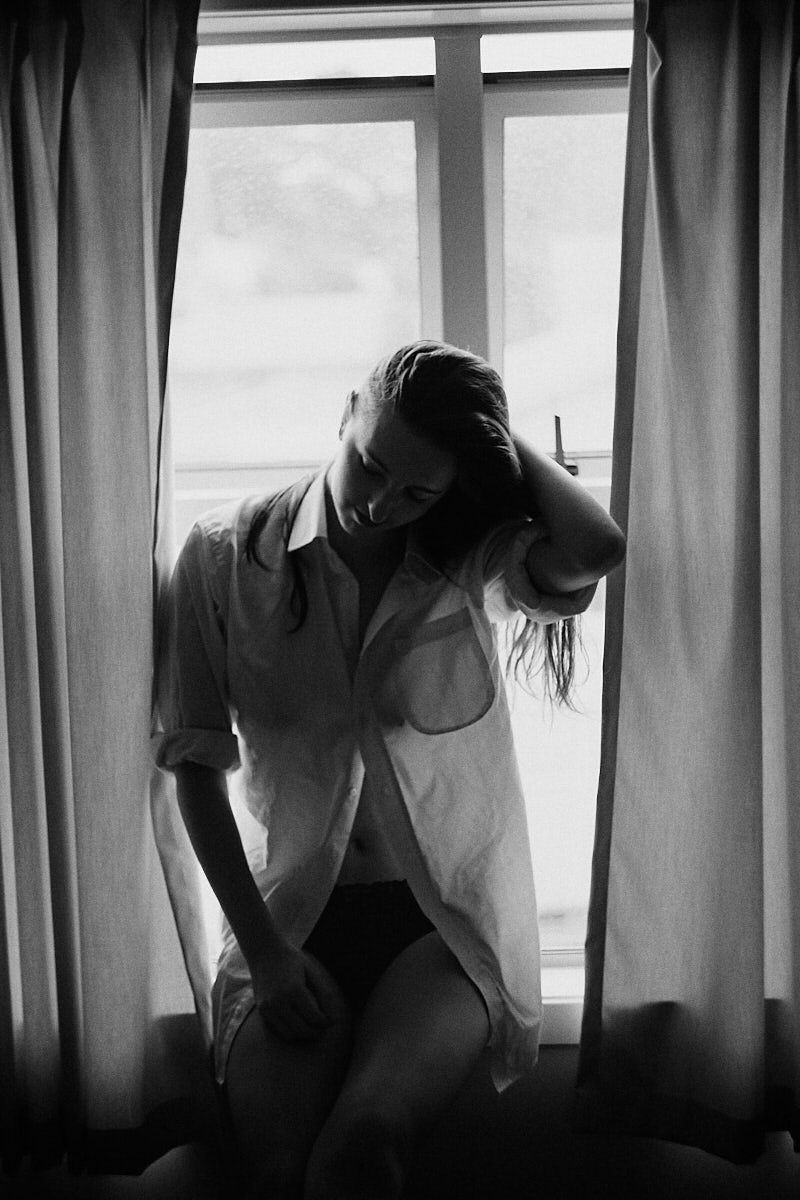 Black and white photo of a woman sitting by a window in an open shirt, hand in hair, soft natural light.