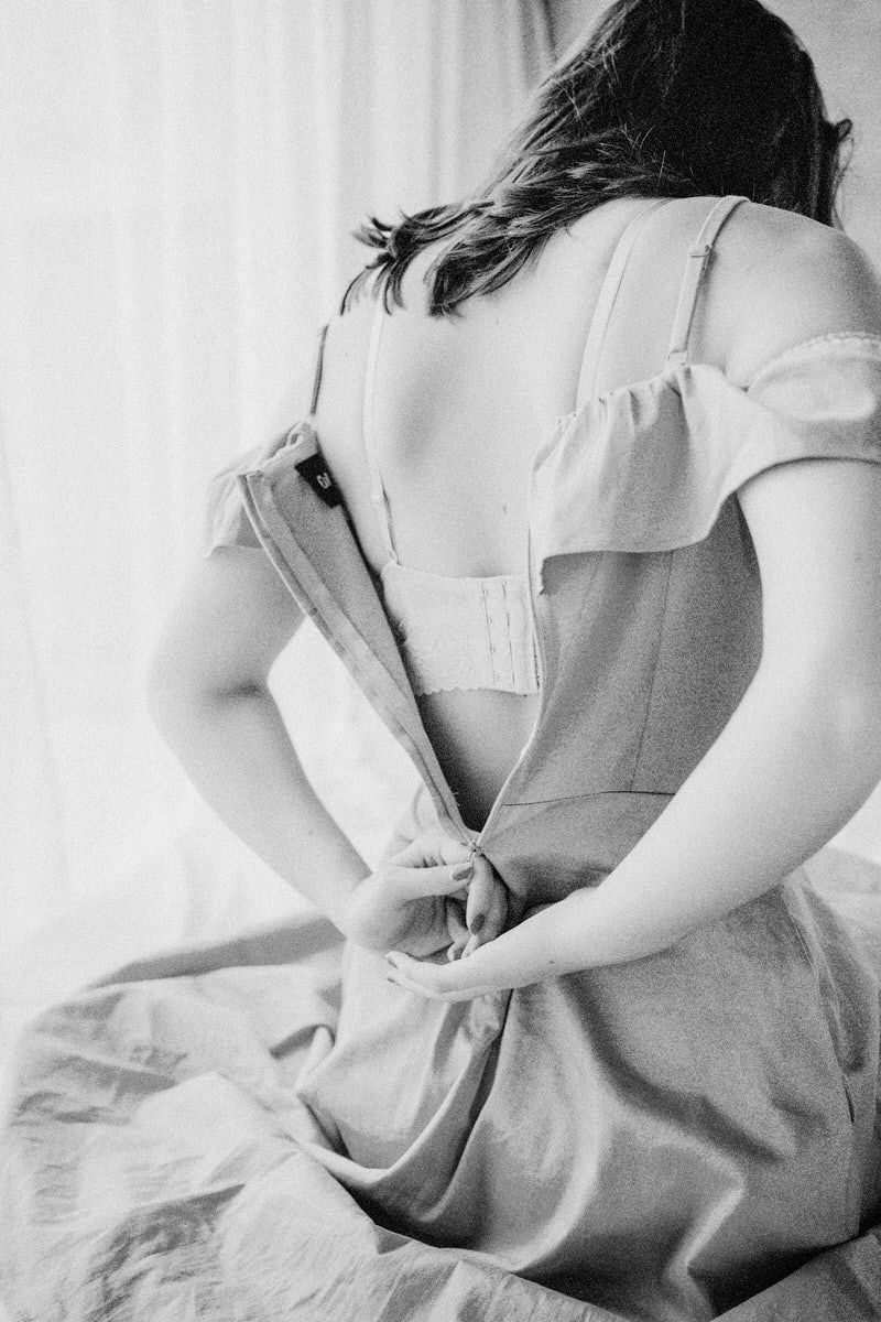 Black and white photo of a woman zipping up the back of her dress while seated near a bright window.