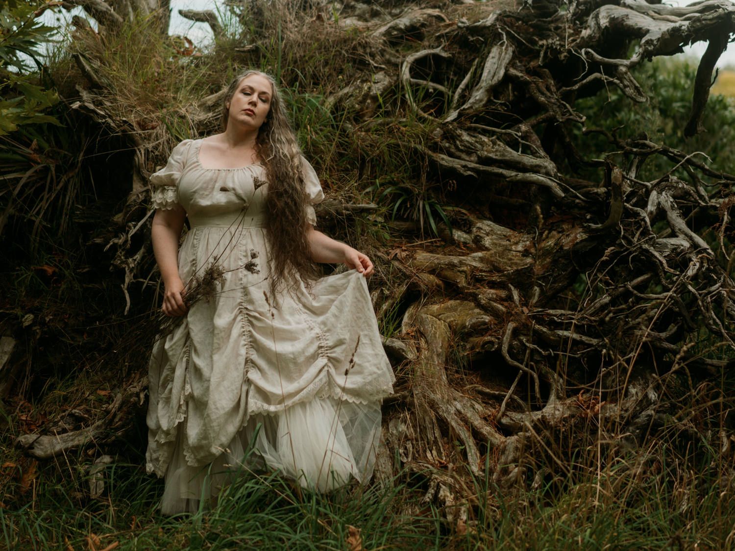 Woman in vintage white gown standing among twisted tree roots in an enchanted forest setting.