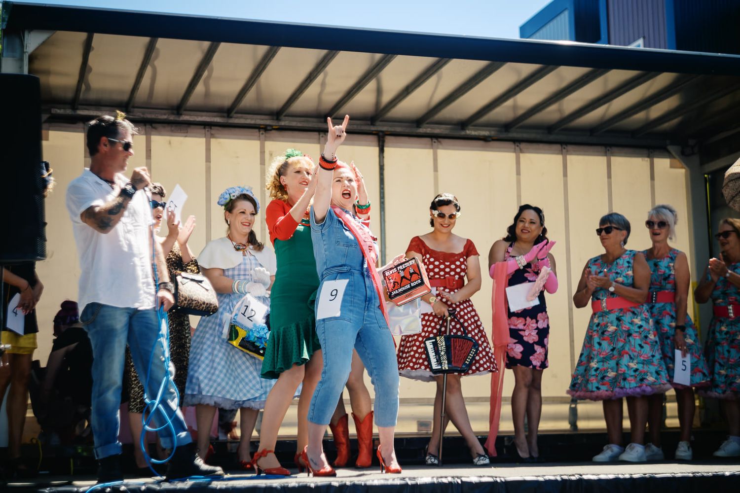 Women in colorful vintage dresses stand on a stage at an outdoor retro-themed event on a sunny day.
