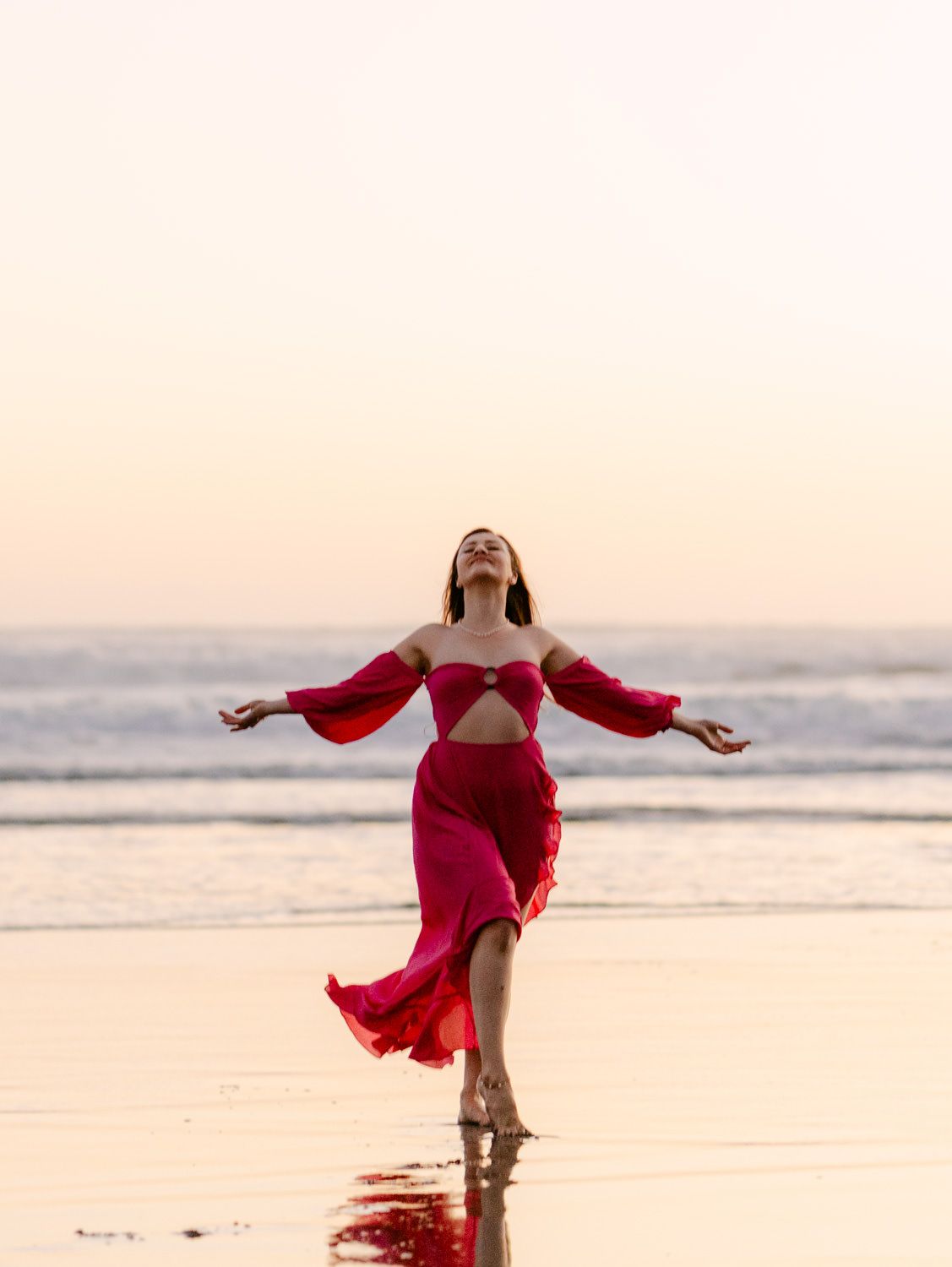 Woman in red dress dancing on beach at sunset with arms outstretched and head tilted back.