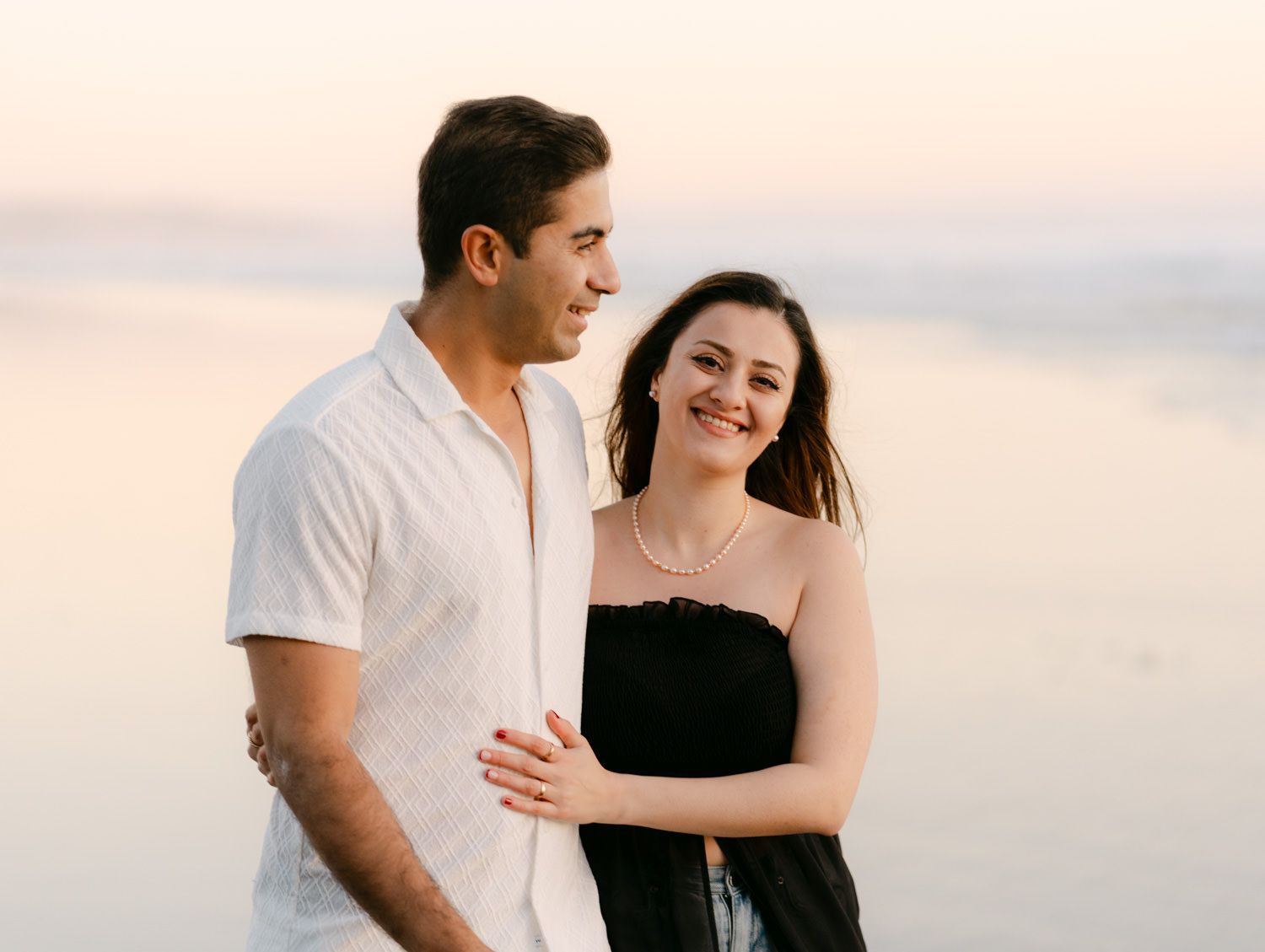 A couple embraces and smiles together on a beach at sunset, the woman in a black top and man in a white shirt.