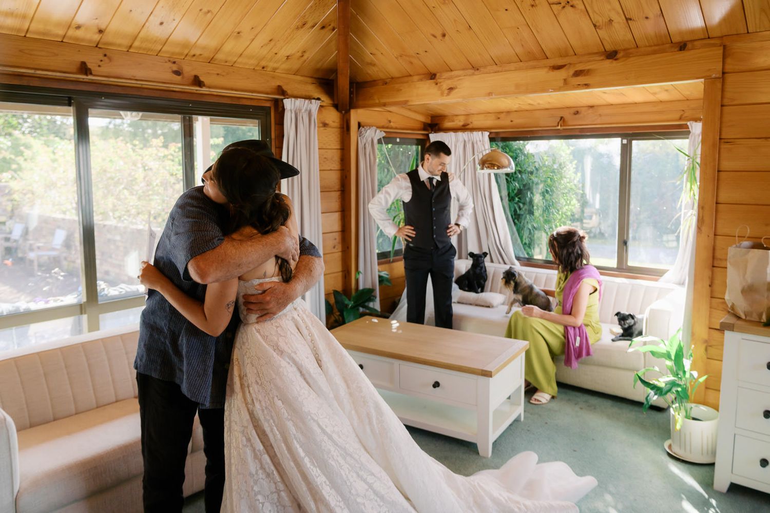 Bride in lace gown embracing guest in rustic wood-paneled wedding venue while groom and bridesmaid look on.