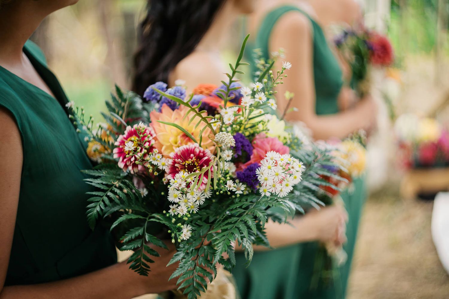 Bridesmaids in emerald green dresses holding vibrant wildflower bouquets with ferns at an outdoor wedding.