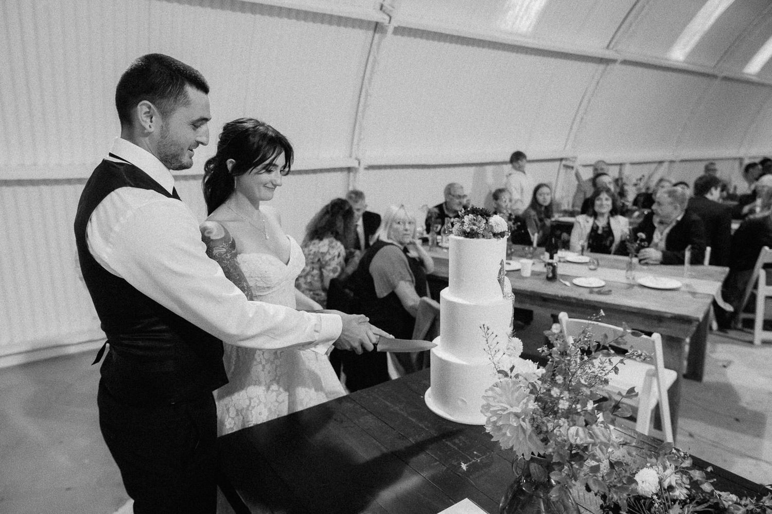 Black and white photo of bride and groom cutting their wedding cake together at a reception tent.