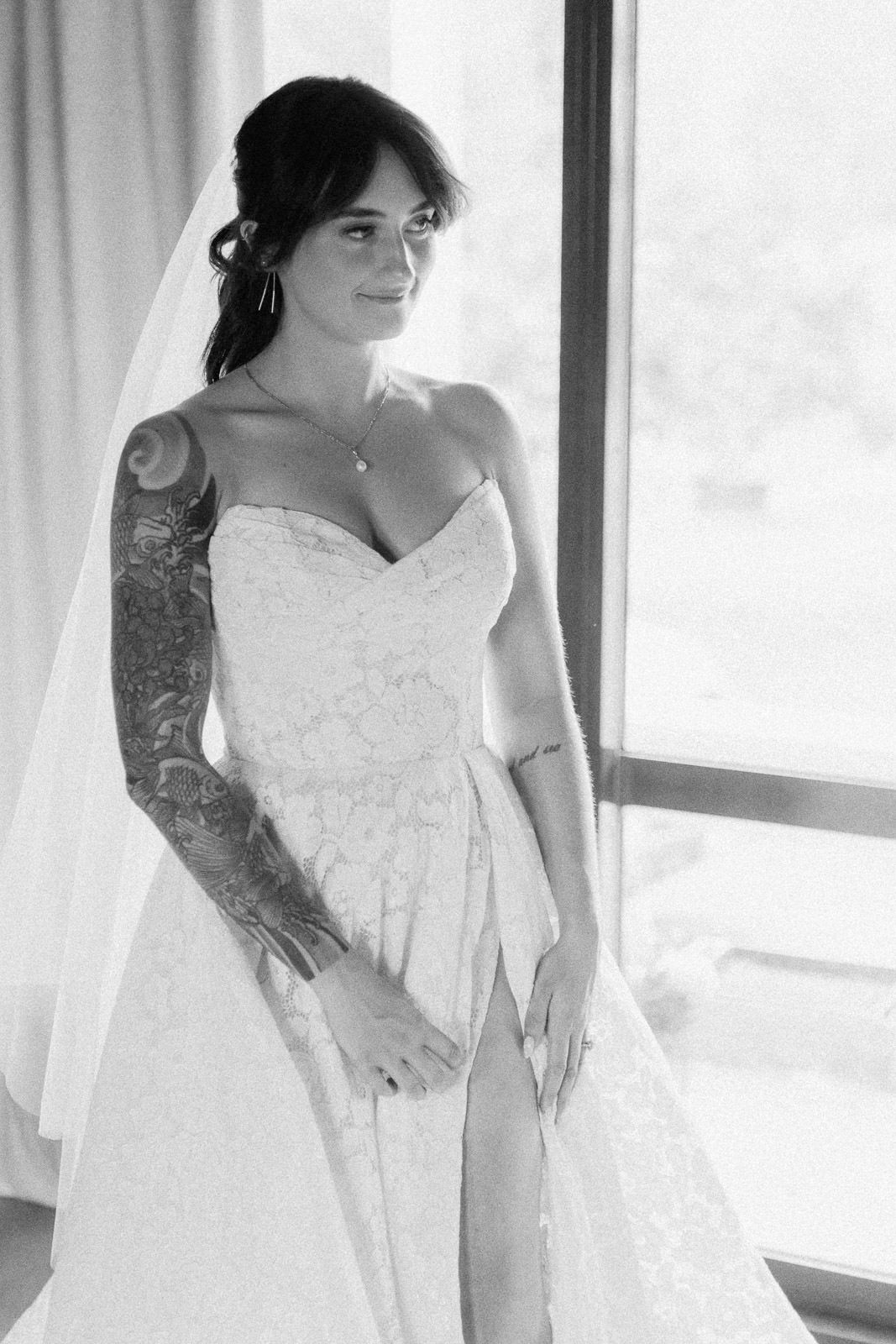 Black and white photo of a tattooed bride in a strapless lace wedding gown with veil, posing near a window.