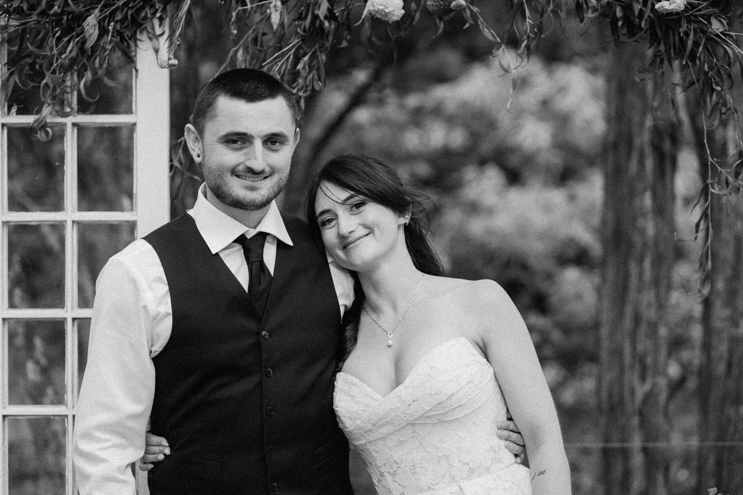 Black and white wedding photo of smiling bride and groom posing together under floral arch outdoors.