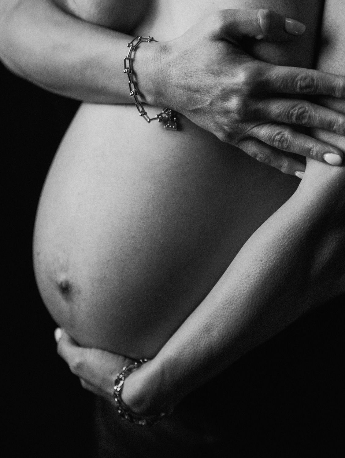 Black and white photo of a pregnant woman cradling her bare baby bump with both hands wearing delicate chain bracelets.