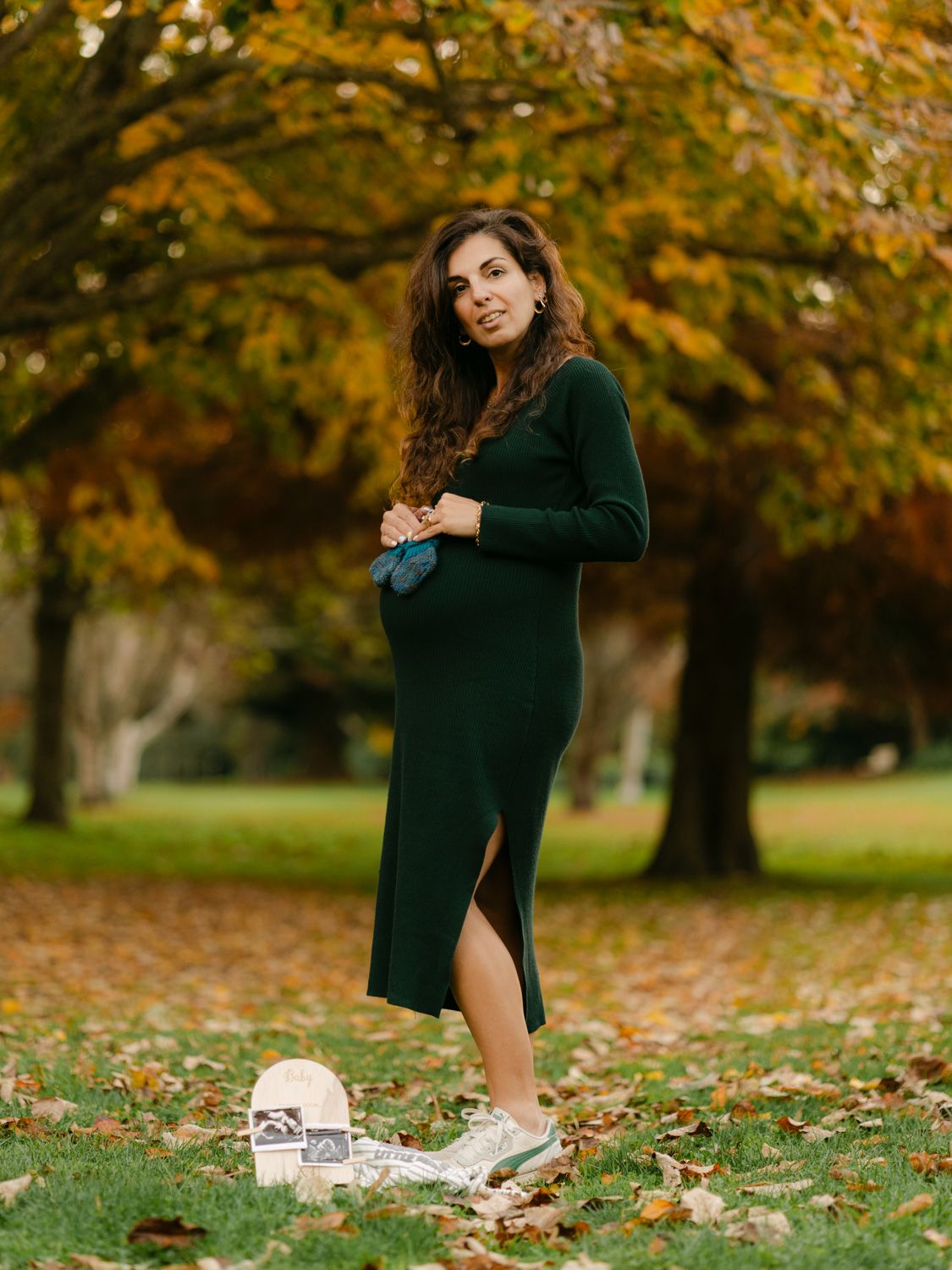 Pregnant woman in green dress holding baby shoes in autumn park with colorful fall foliage.