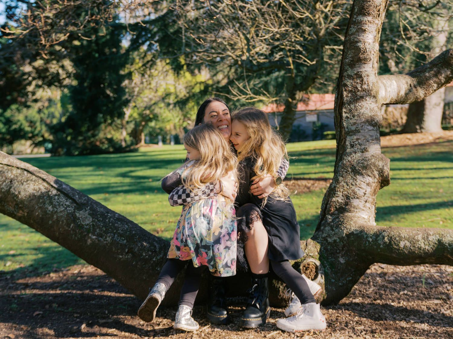A woman and two young girls share a warm hug while sitting on a large tree branch in a green park.