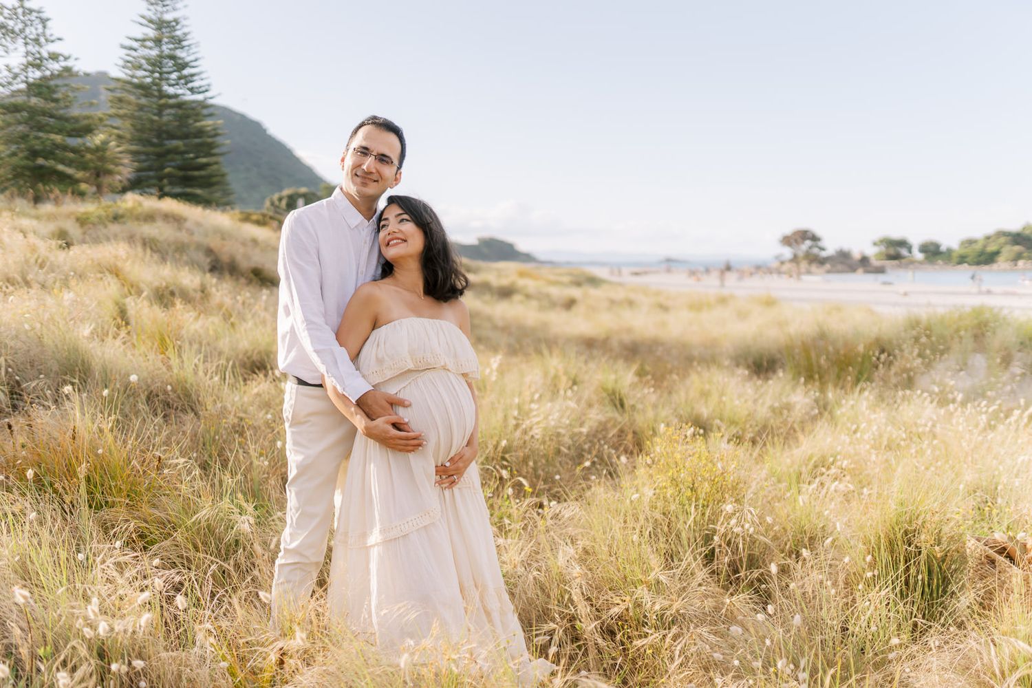 Couple in a golden field, pregnant woman in white dress, man embracing her from behind, outdoor maternity photo.