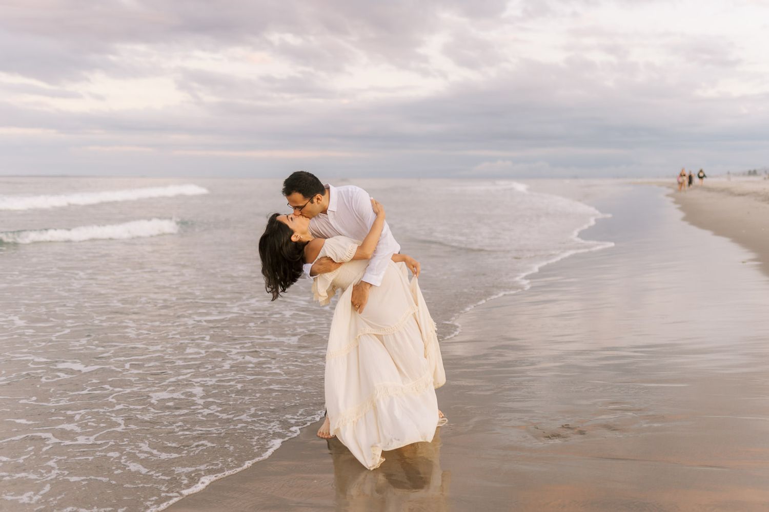 Romantic couple sharing a dip kiss on a sandy beach with ocean waves behind them.